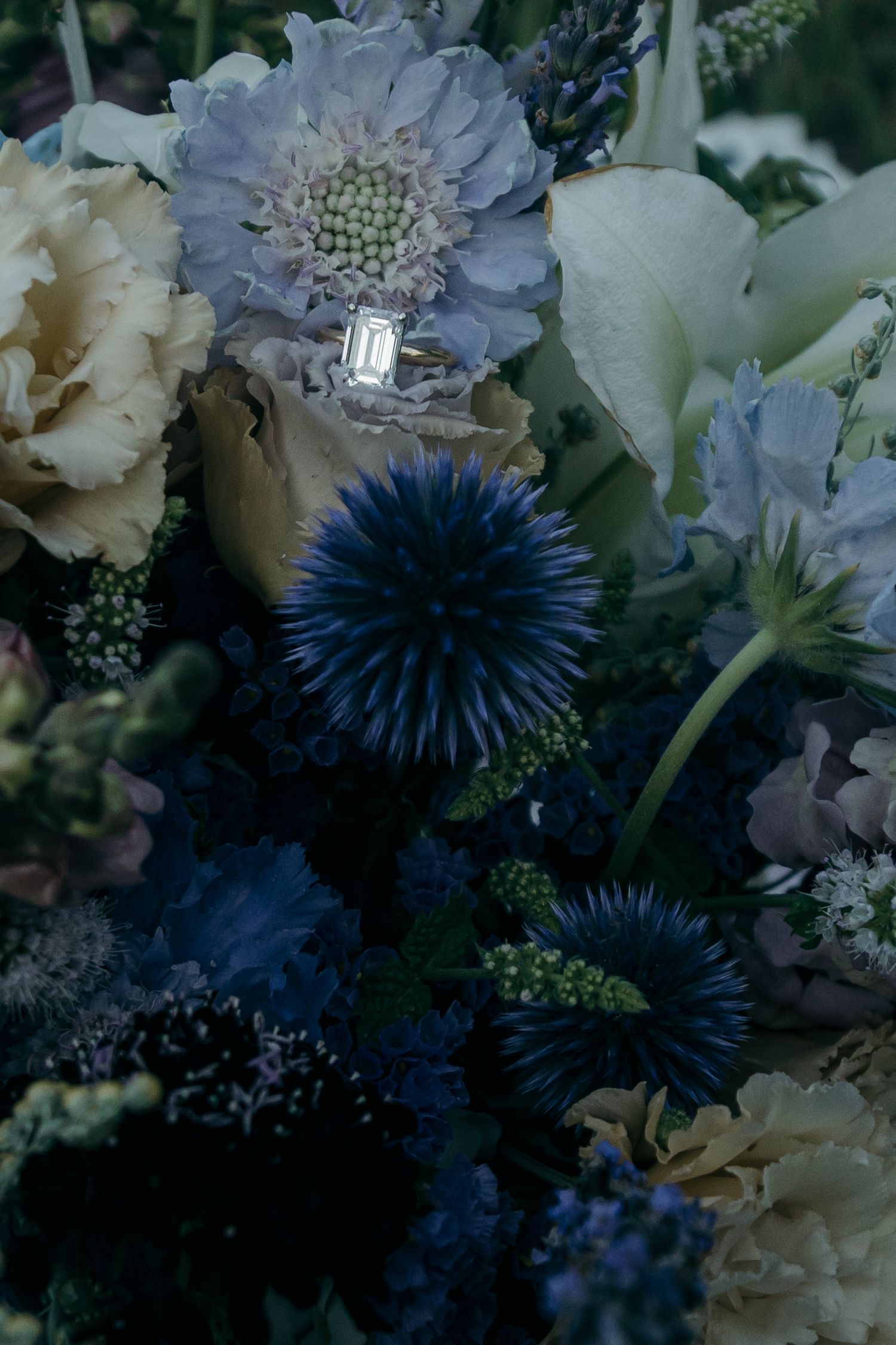 Close-up of emerald cut engagement ring nestled among blue and cream wildflowers in bridal bouquet.