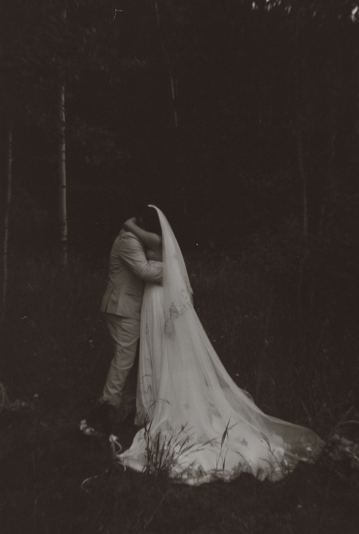 Romantic black and white photo of bride and groom kissing in dark forest, brides cathedral veil trailing on ground.