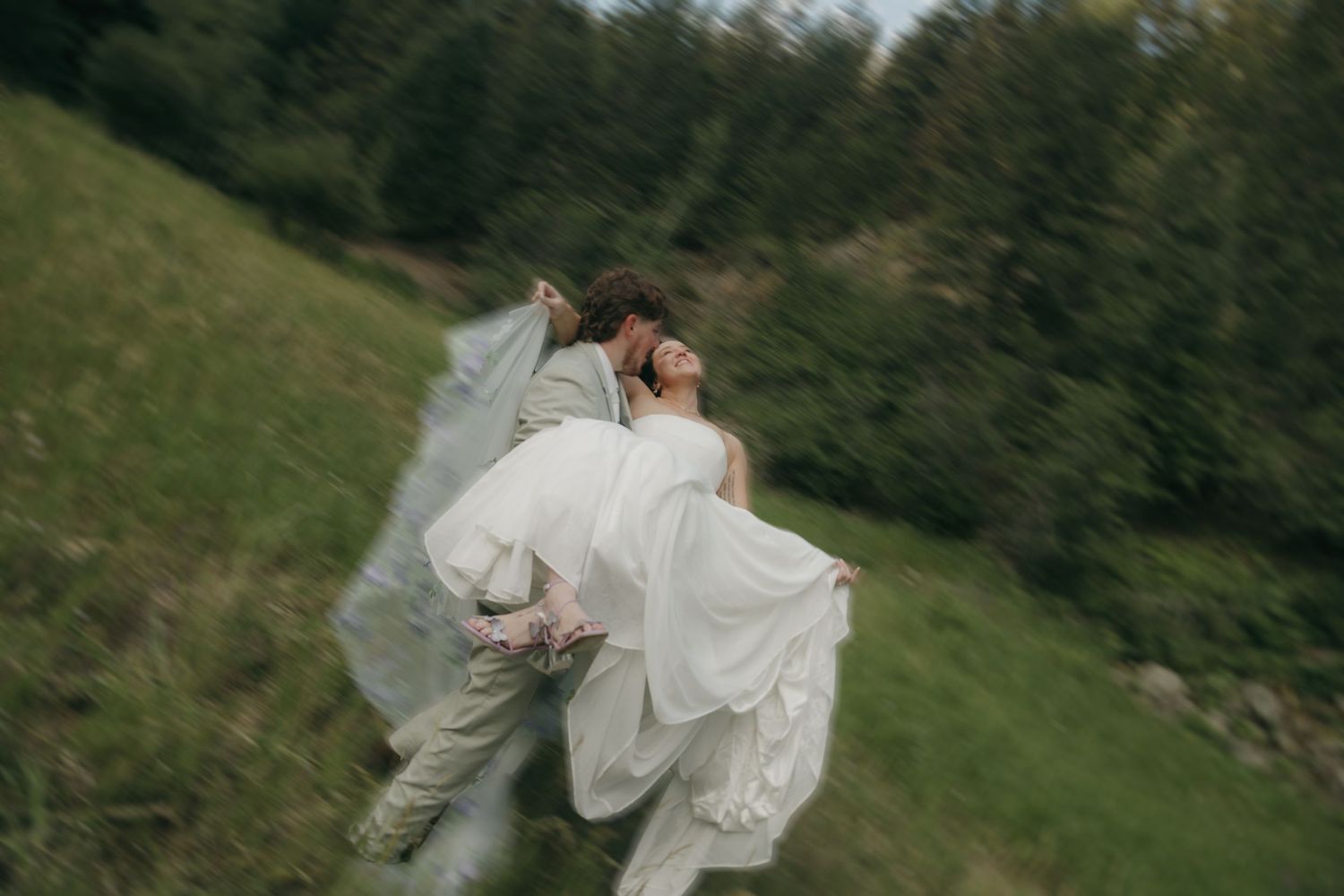 Groom spins bride in white gown across green mountain meadow in joyful motion-blur wedding photo.