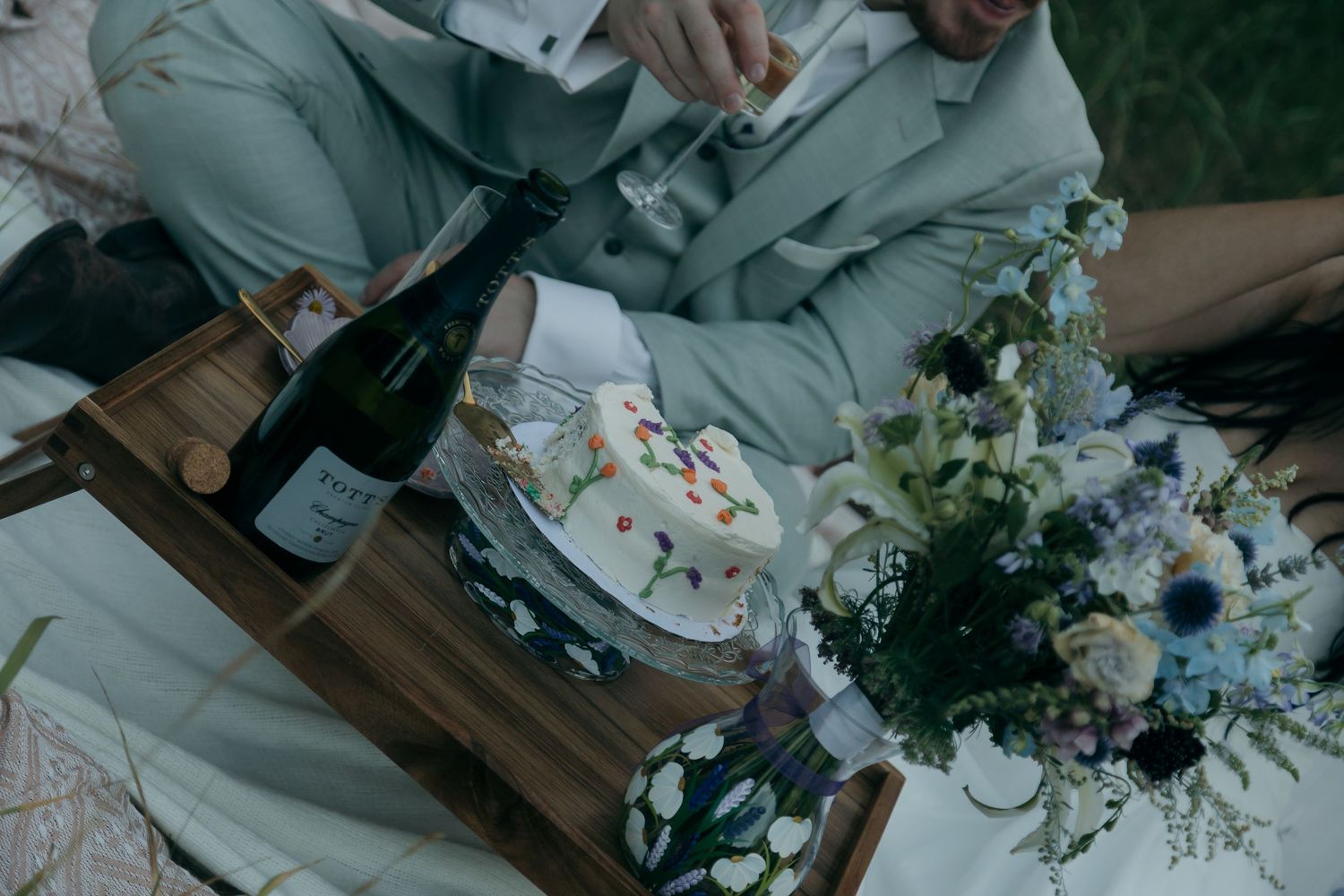 Overhead view of wedding couple with champagne bottle, floral cake, and blue bouquet on a picnic blanket.