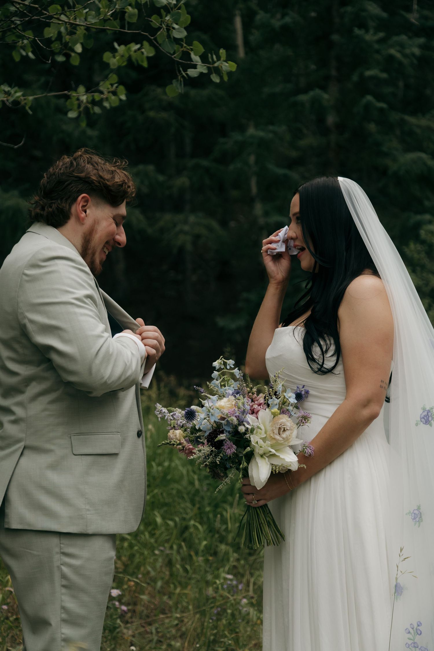 Emotional bride and groom cry tears of joy during forest wedding ceremony, bride holding blue wildflower bouquet.