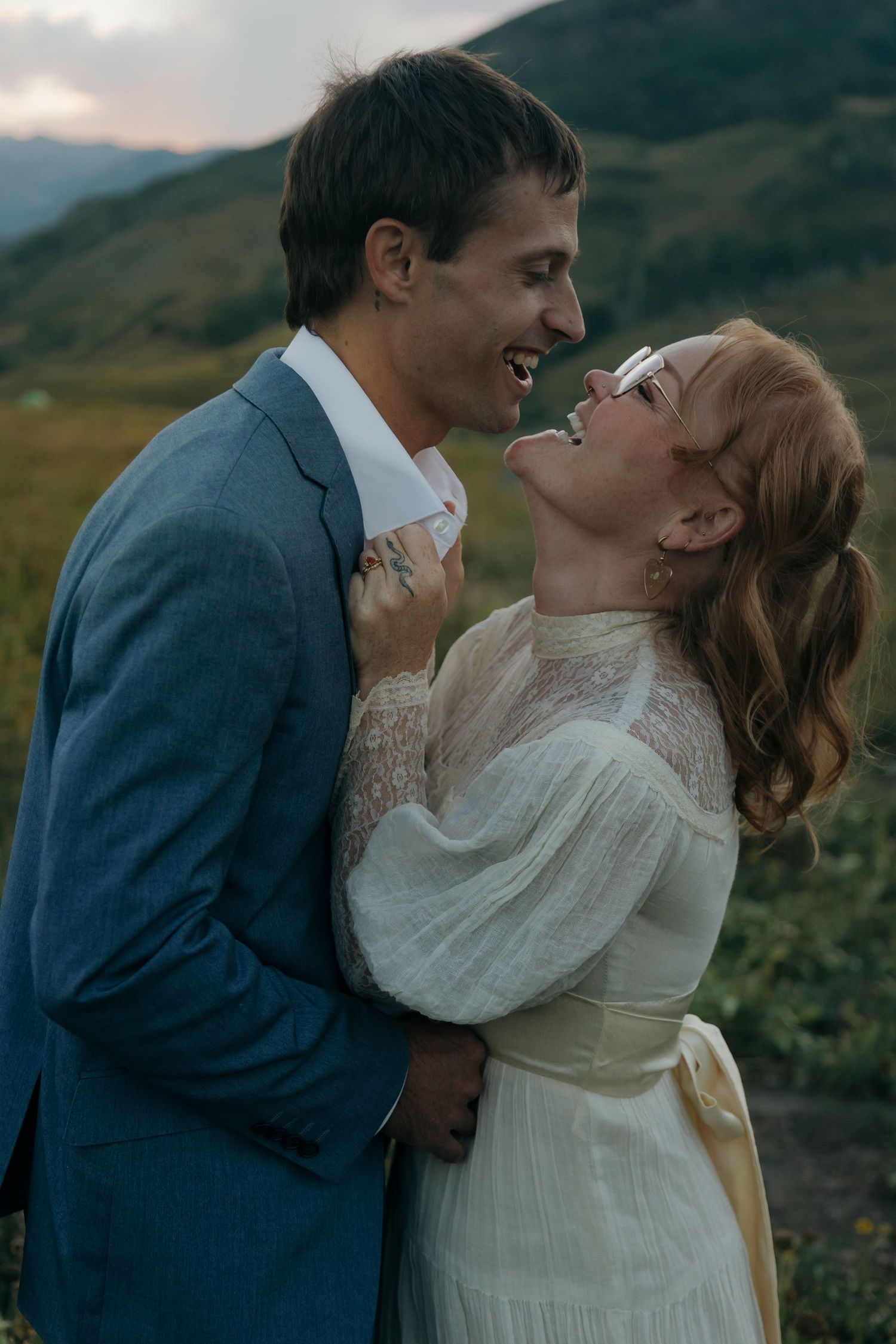 Laughing bride and groom embrace outdoors with mountain backdrop during wedding celebration.