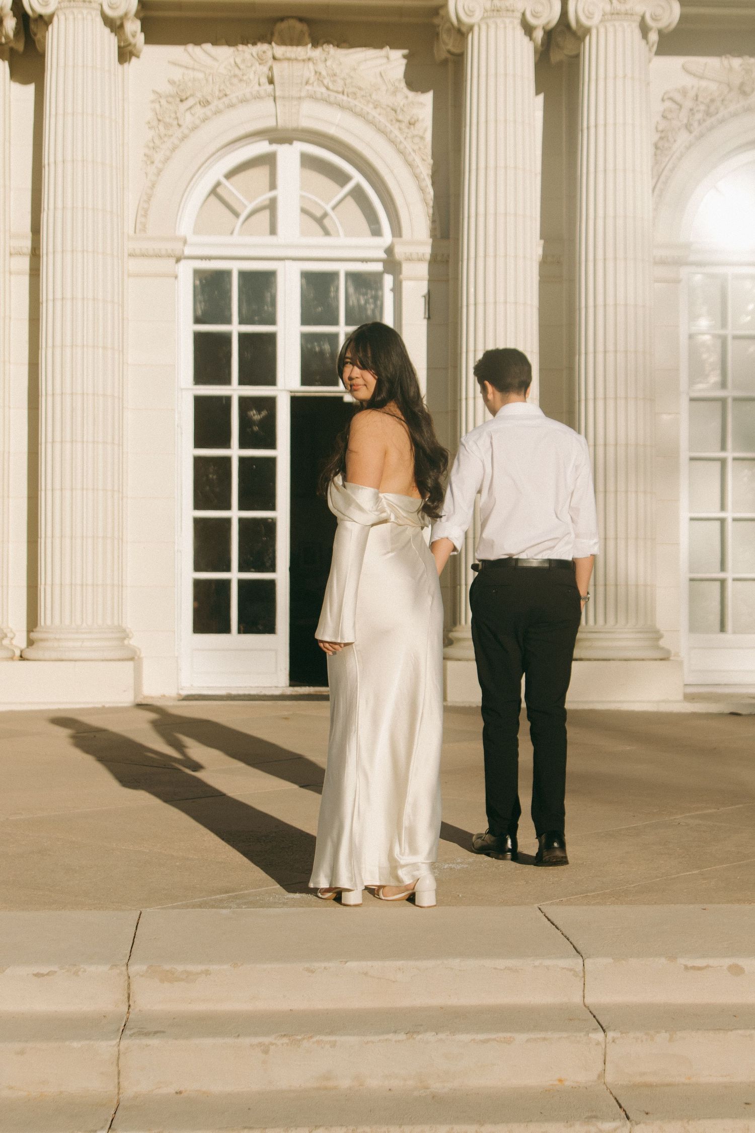 A couple walking along a classical building exterior with white columns and arched windows.