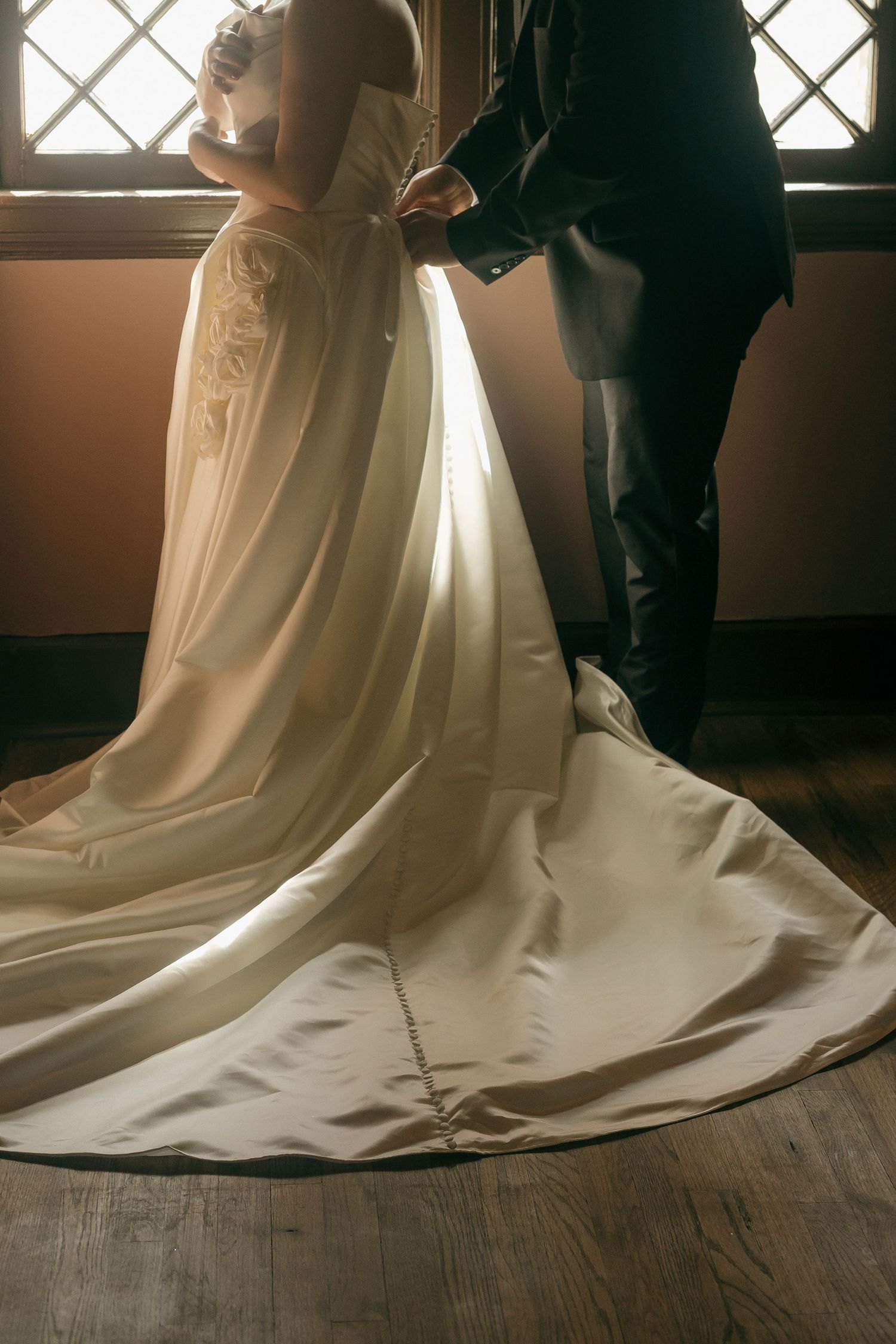 Close-up detail of a flowing white wedding dress train and formal attire near a window.
