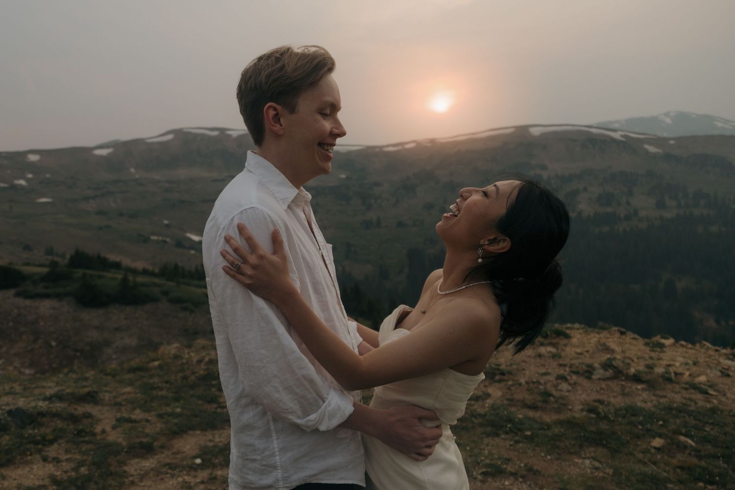 Couple embraces at golden sunset on mountain overlook, bride in white dress gazing up at groom.