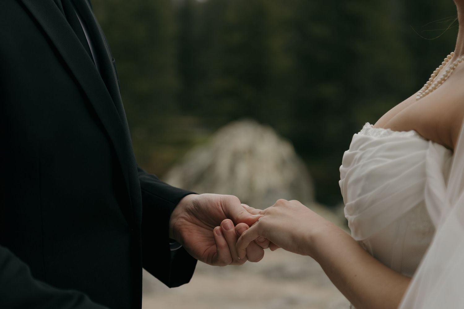 Close-up of bride and groom holding hands, her in white lace gown, him in dark suit, near a forest.