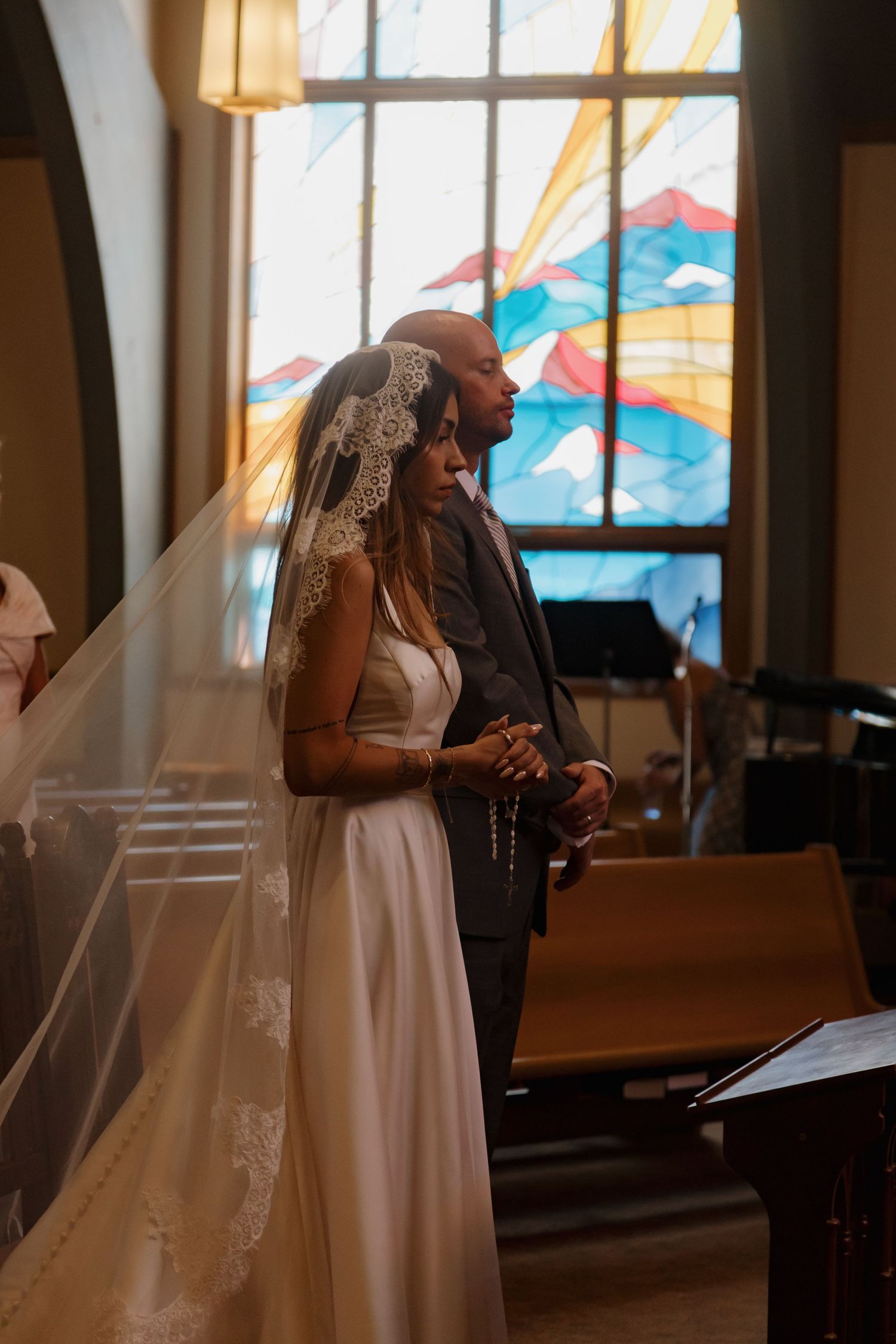 A bride in a lace veil and groom pray together before a vibrant stained glass window.