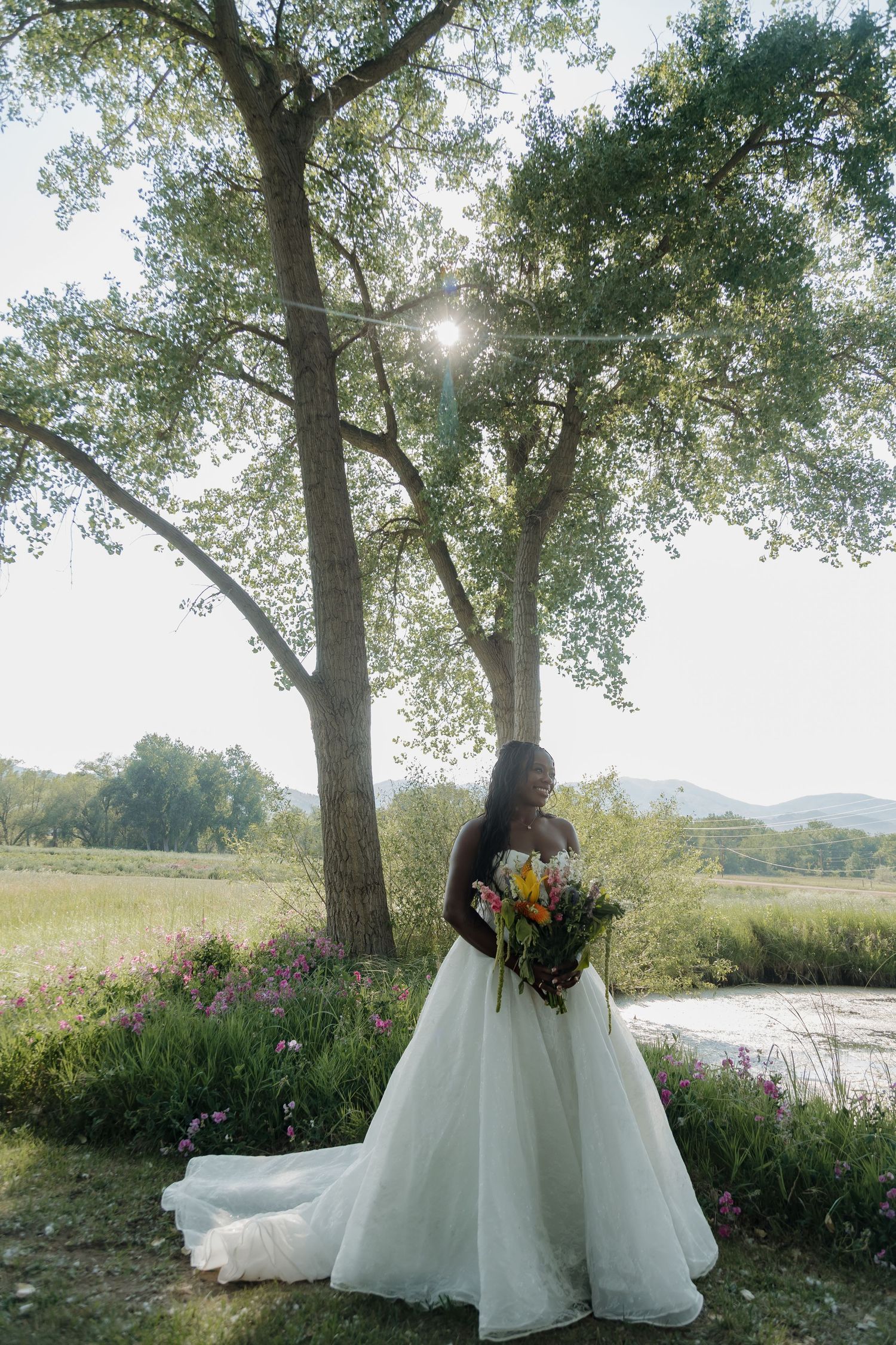 Black bride in a ball gown holding a colorful bouquet standing by trees near a scenic meadow.