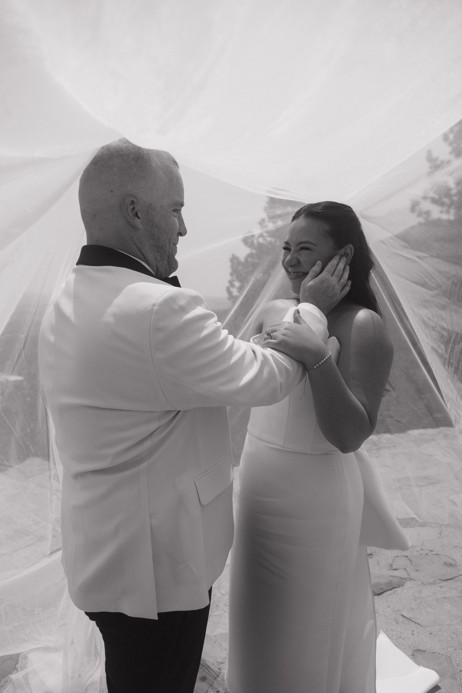 Black and white photo of groom tenderly cupping brides face under her flowing veil at an outdoor wedding ceremony.