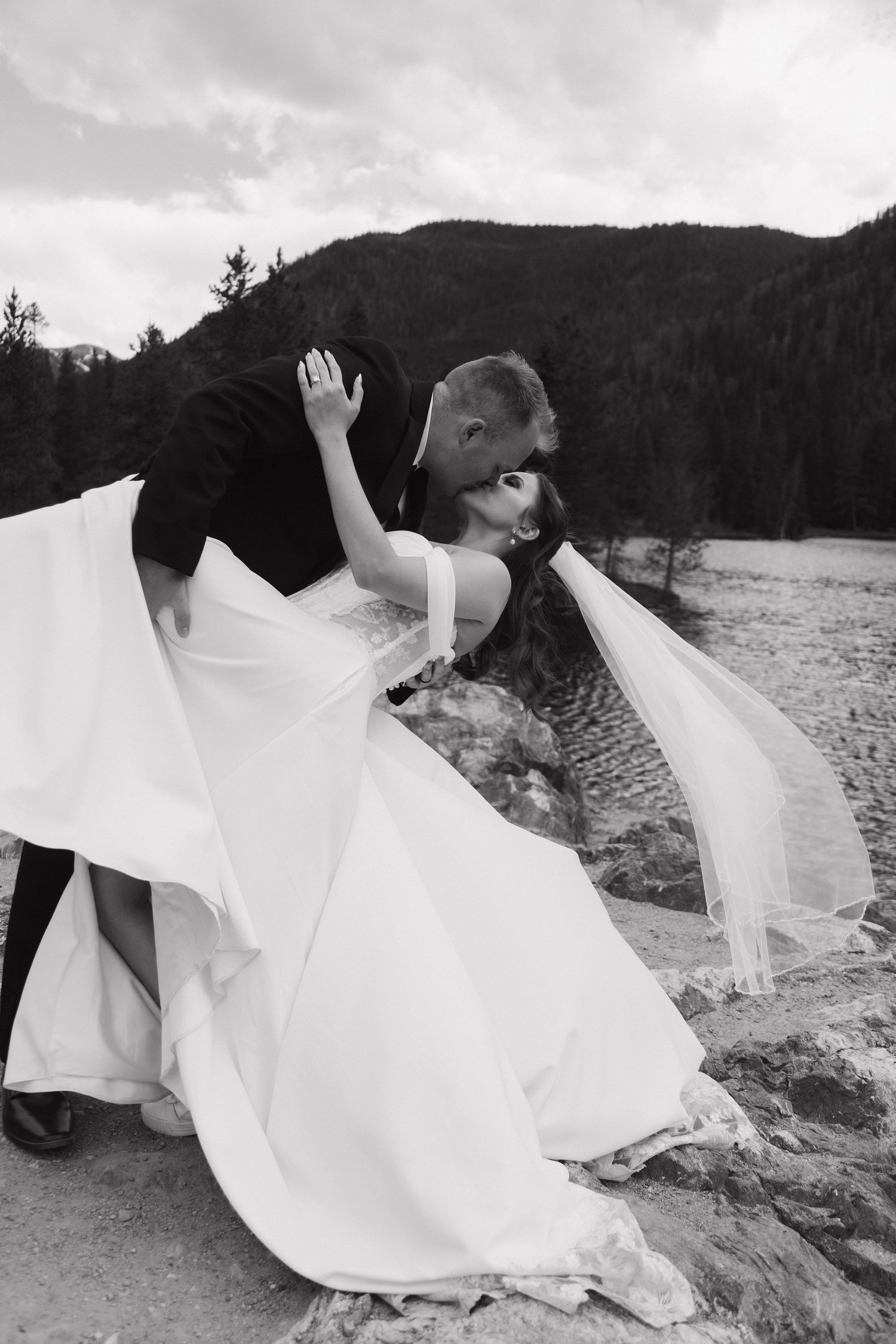 Black and white photo of a groom dipping his bride for a kiss outdoors with mountain forest backdrop.