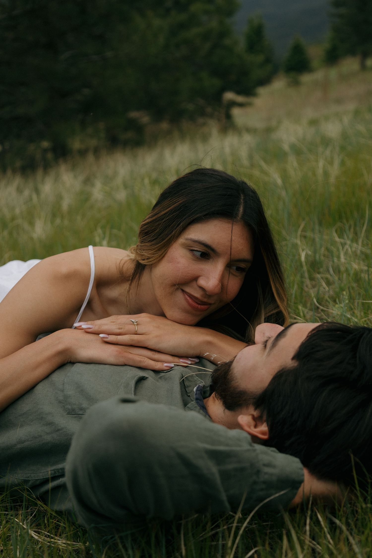 A couple lying in a grassy meadow, sharing a tender moment with mountains in the background.