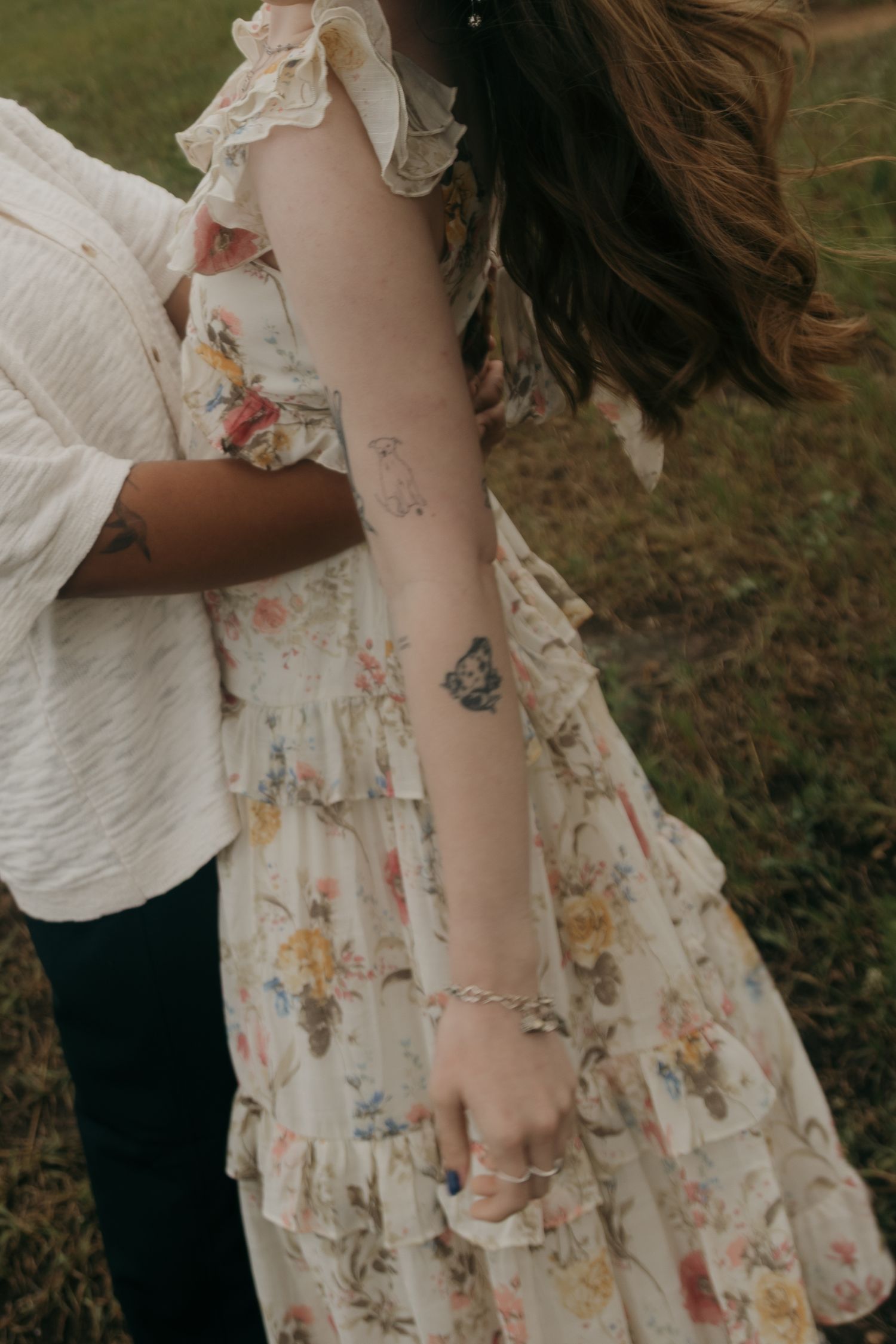Close-up of a woman in a floral dress with butterfly tattoo on her forearm, embraced by a partner outdoors.