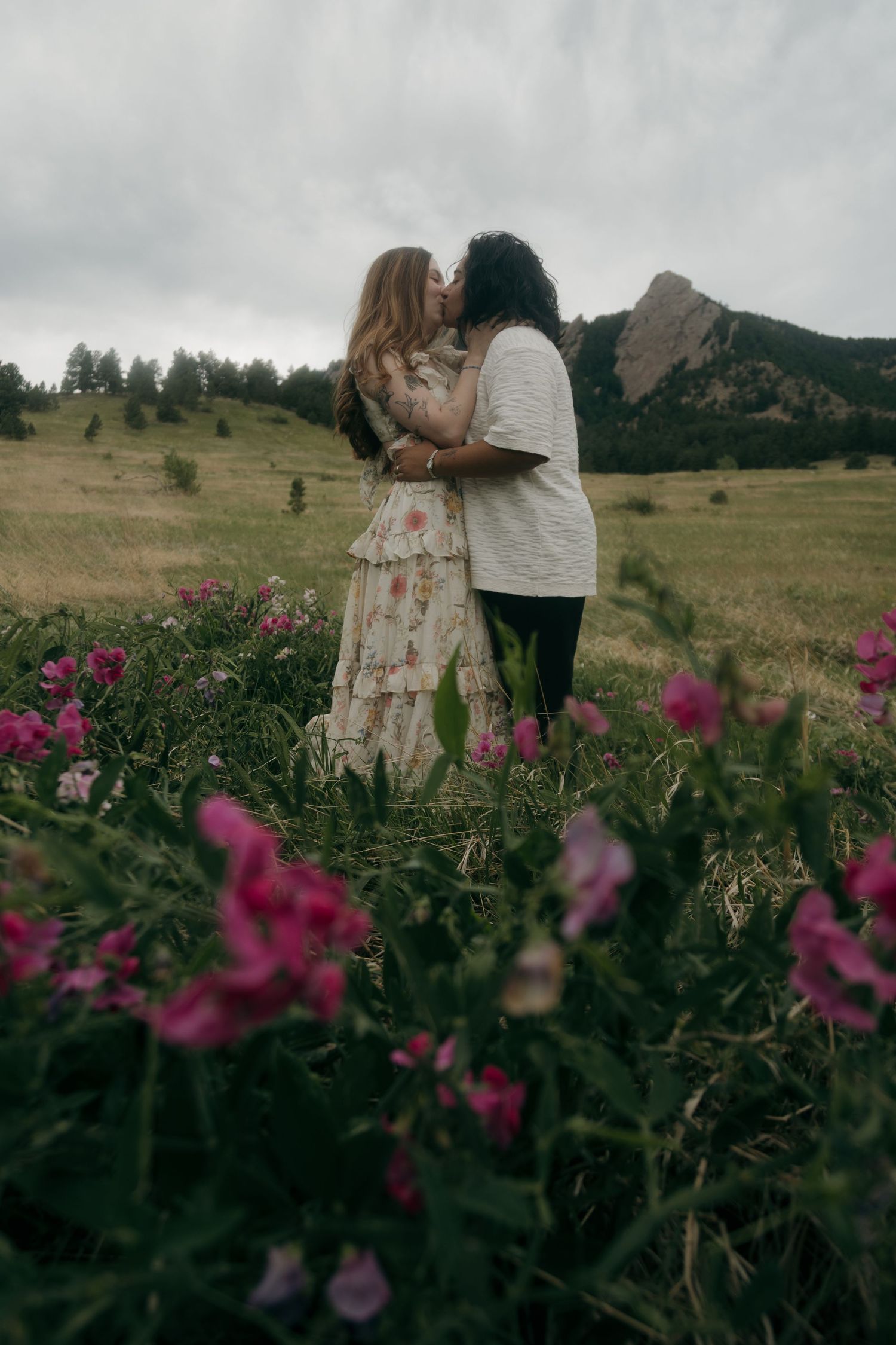 Couple sharing a kiss surrounded by pink wildflowers with the Flatirons mountains visible in the background.