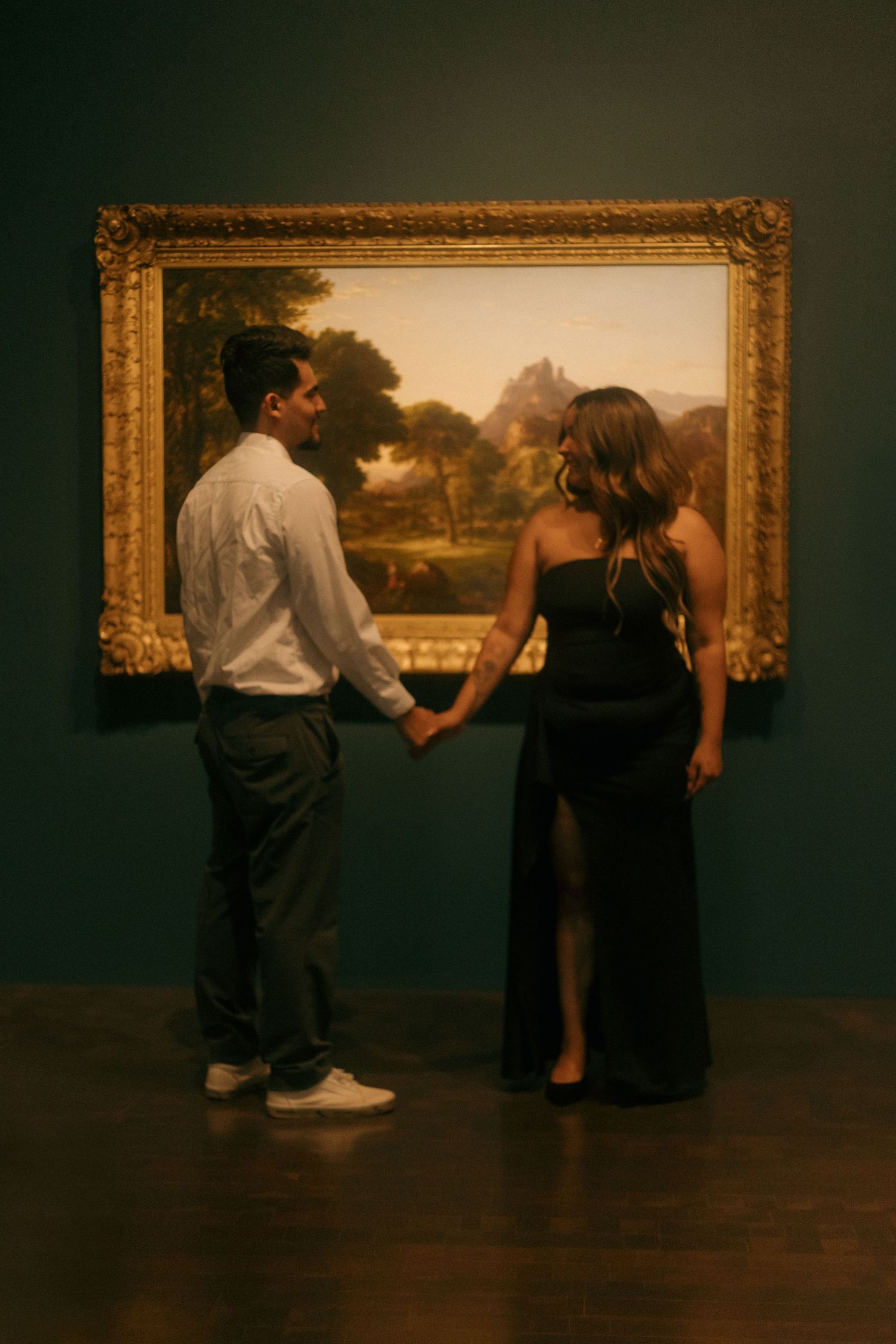 Couple holding hands in art museum before large gold-framed landscape painting on teal wall.