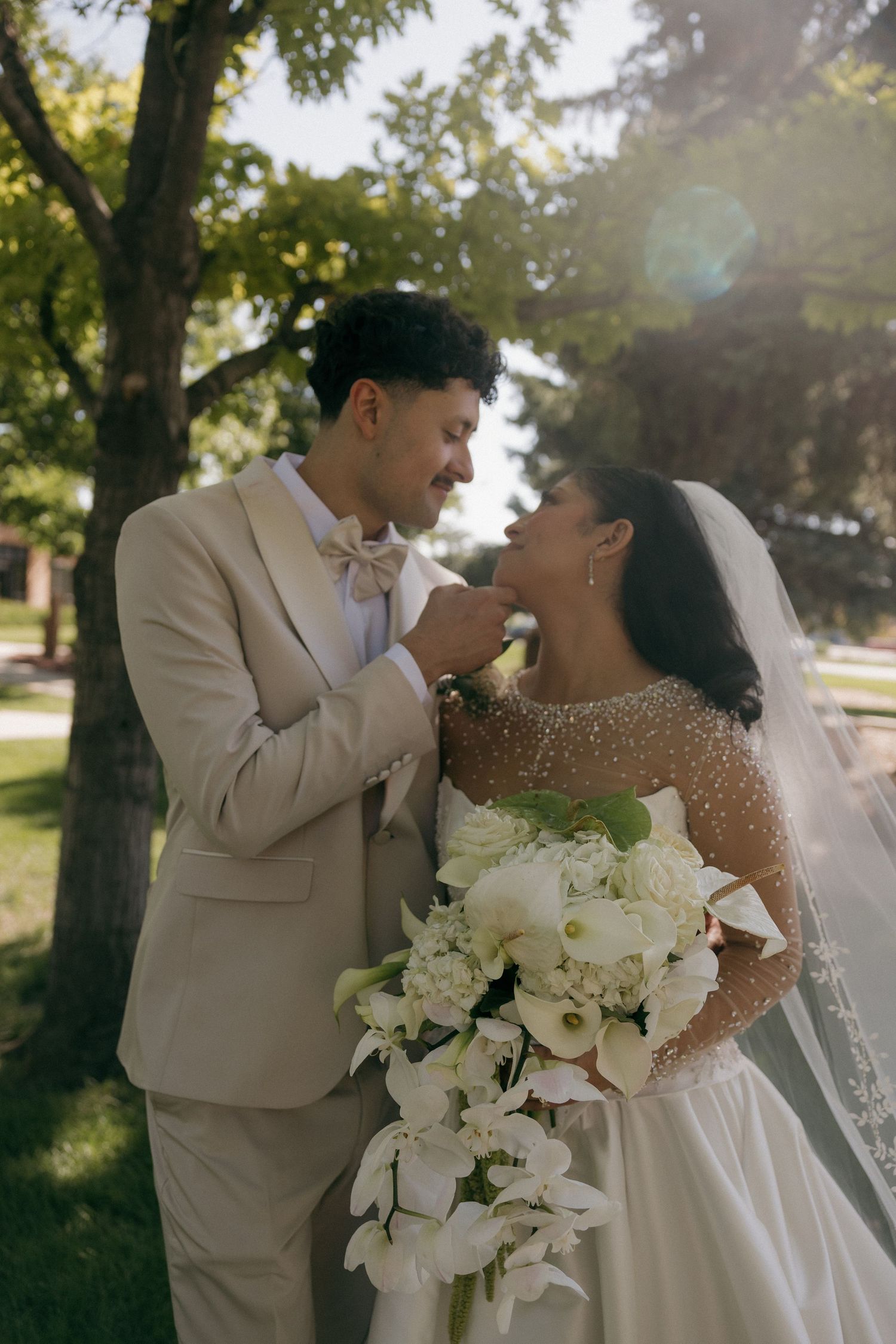 A groom in a beige suit tenderly touches his bride's chin as she holds white flowers outdoors.