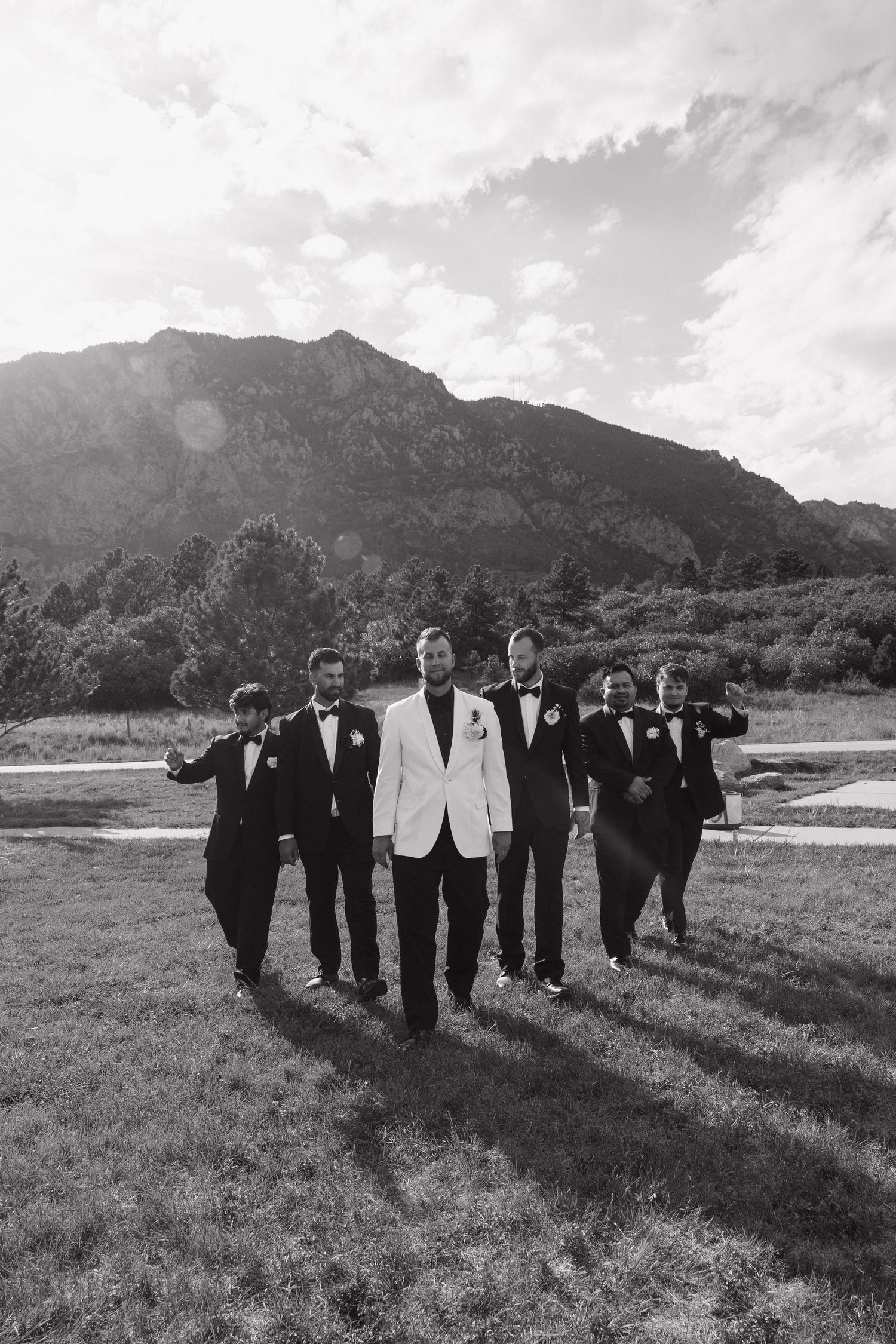 Groom in white jacket walks with groomsmen in black suits before mountain backdrop.