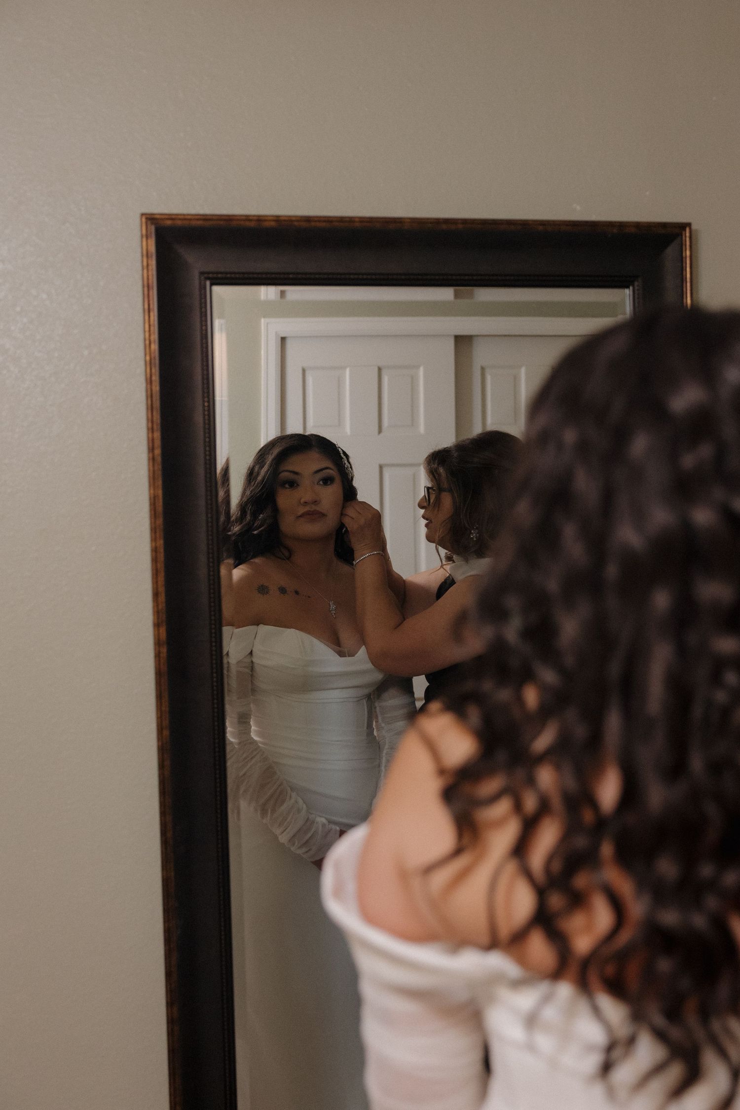 Bride getting earrings adjusted by bridesmaid while wearing white strapless wedding gown, reflected in mirror.