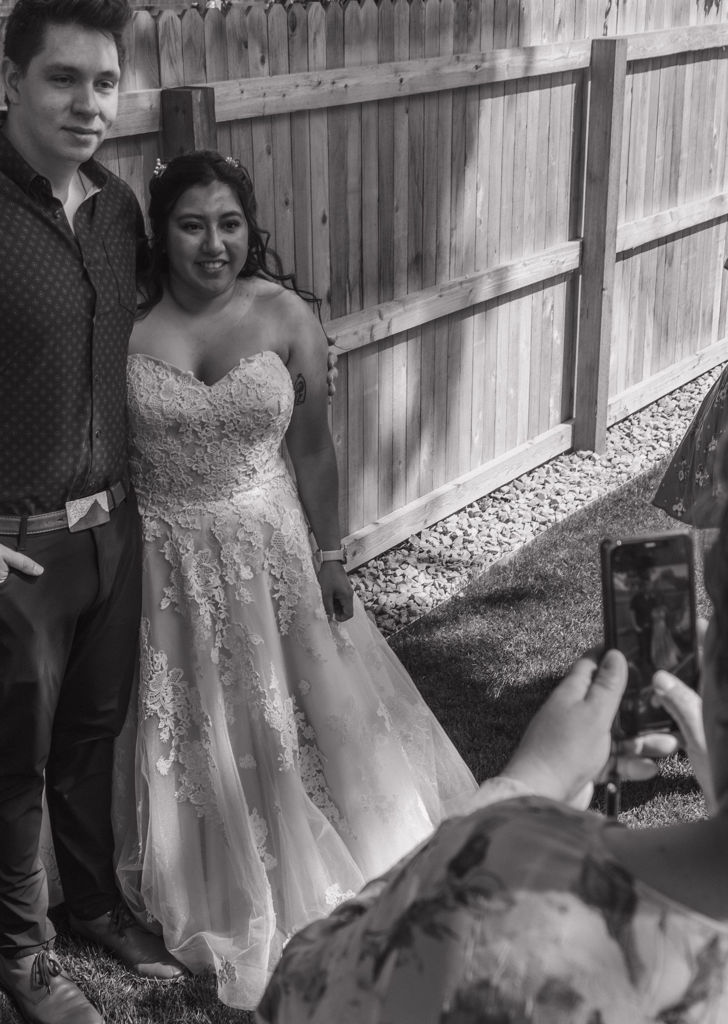 Couple poses together while looking down at the bride's flowing white wedding dress in front of a wooden fence.