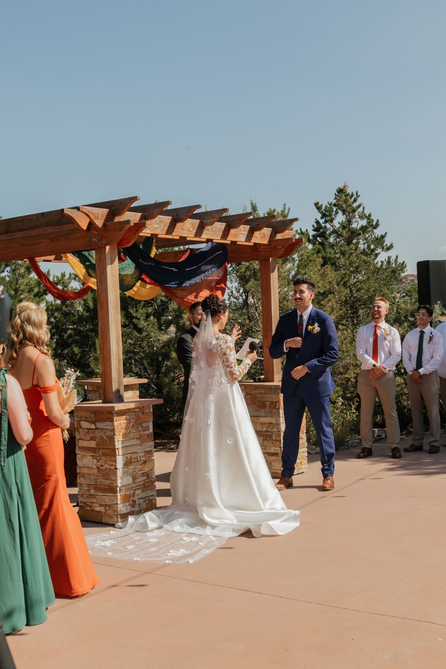 Outdoor wedding ceremony under a wooden pergola with mountain views and bridesmaids in orange and mint dresses.