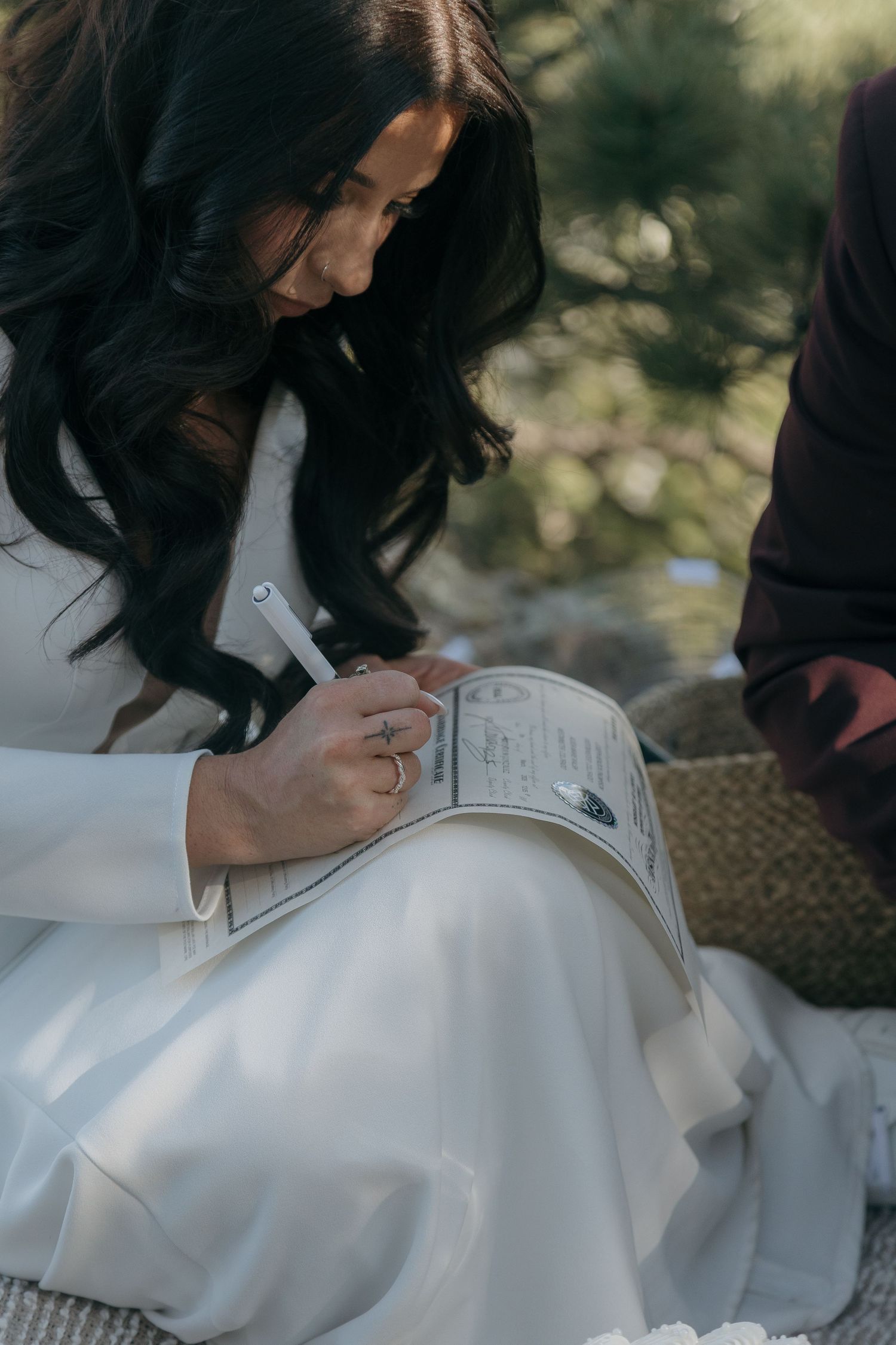 Bride in white gown signs marriage license document while seated outdoors among pine trees.