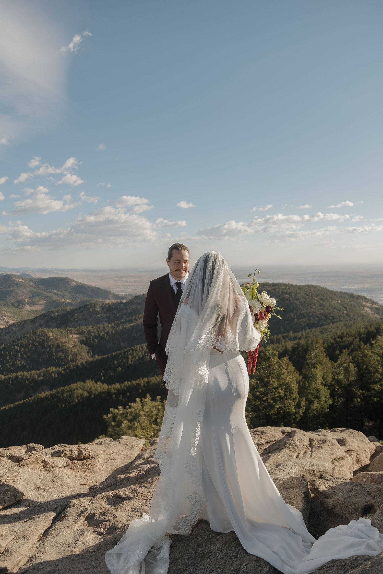 Bride holding bouquet and groom in burgundy suit stand on rocky mountain summit with scenic valley backdrop.