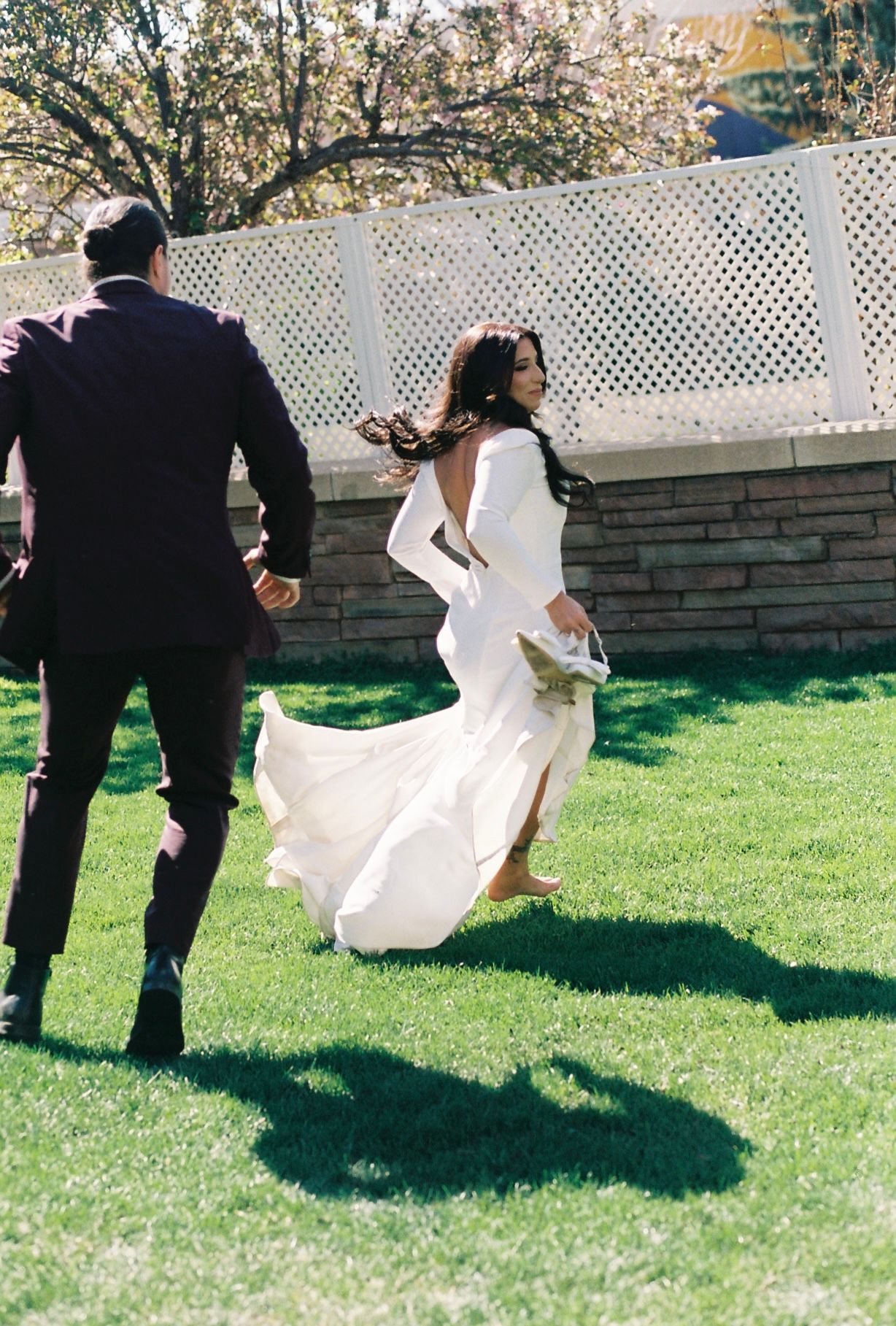 Bride in flowing white gown runs hand-in-hand with groom across green lawn on sunny wedding day.