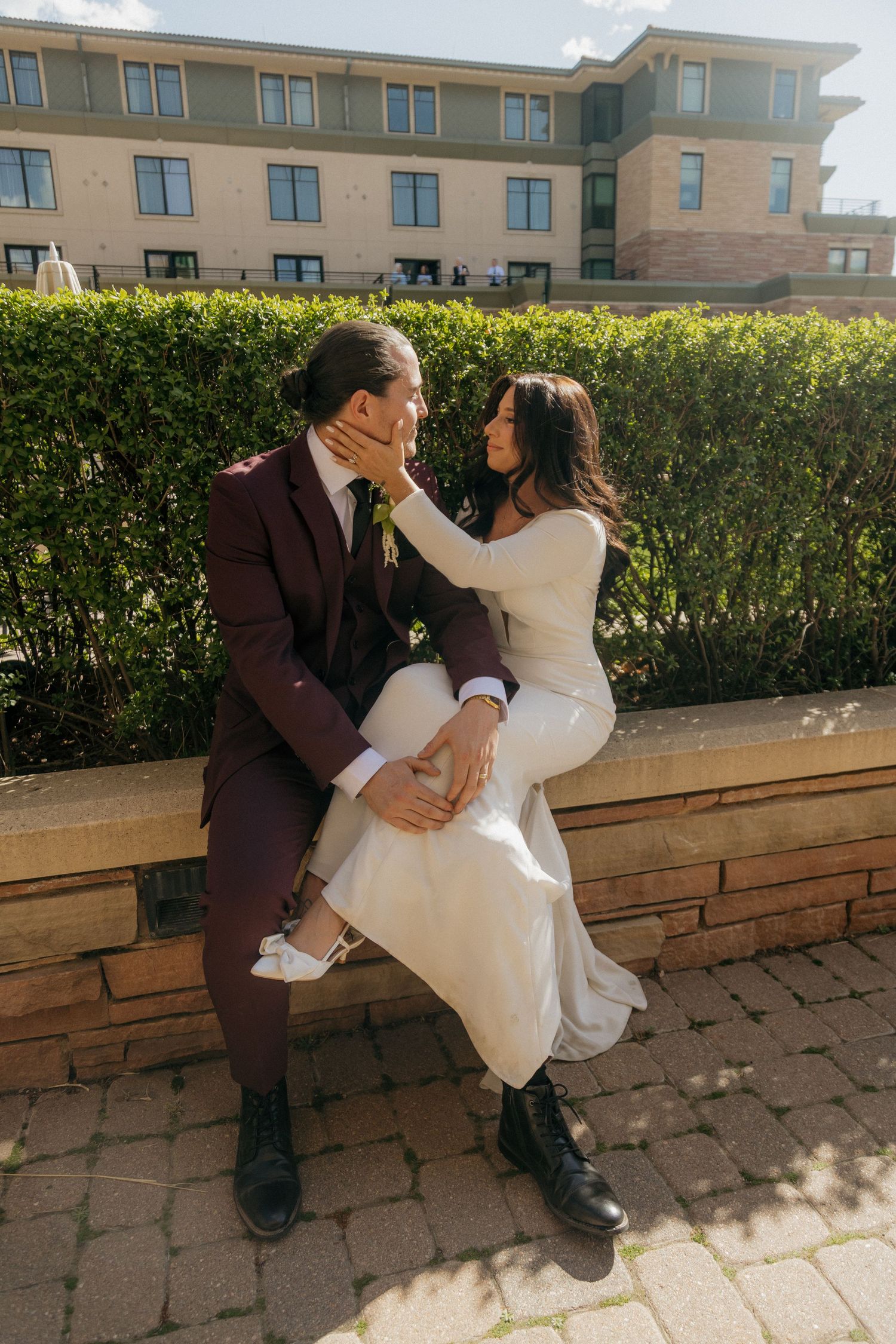 Bride in white gown tenderly touching grooms face while sitting together on a stone ledge in sunlight.
