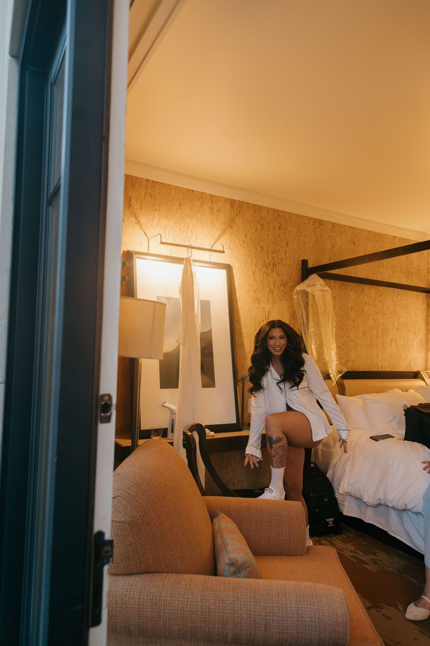 A bride in a white robe stands in a warmly lit hotel room preparing for her wedding day.