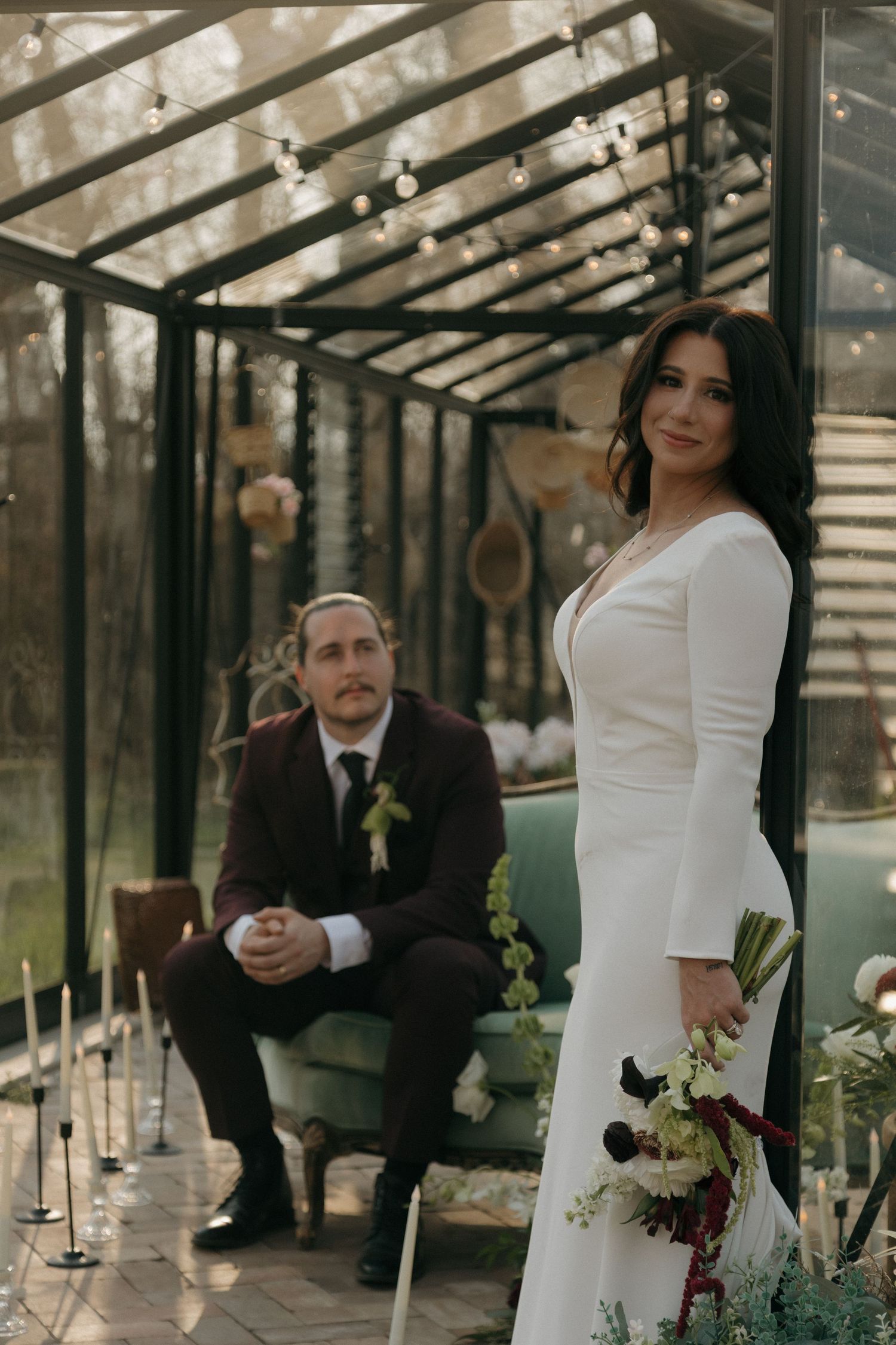 Bride in long-sleeve white gown holds bouquet while groom in burgundy suit sits in greenhouse.