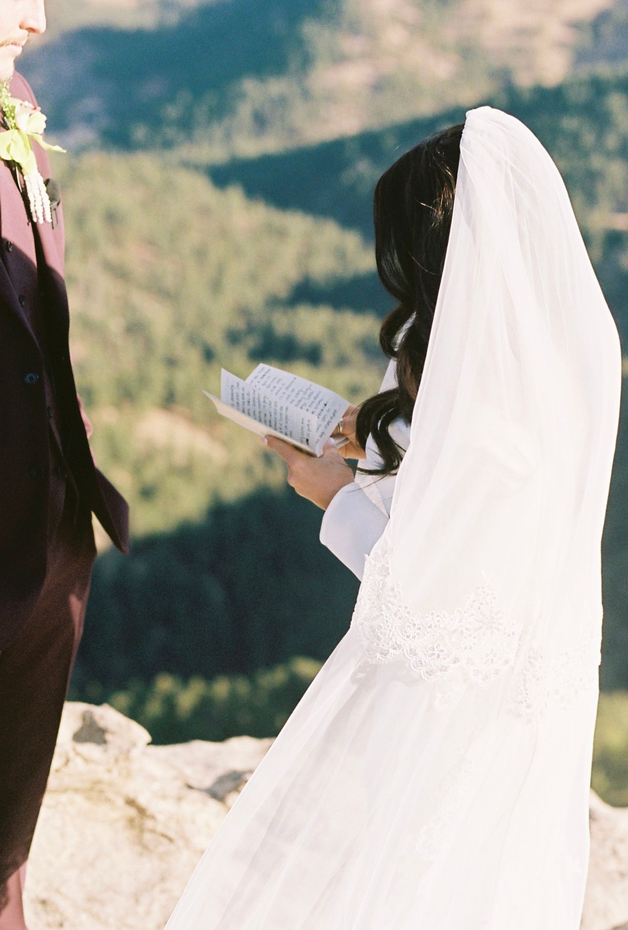 Bride in white lace gown and veil reads wedding vows from book on mountain overlook.