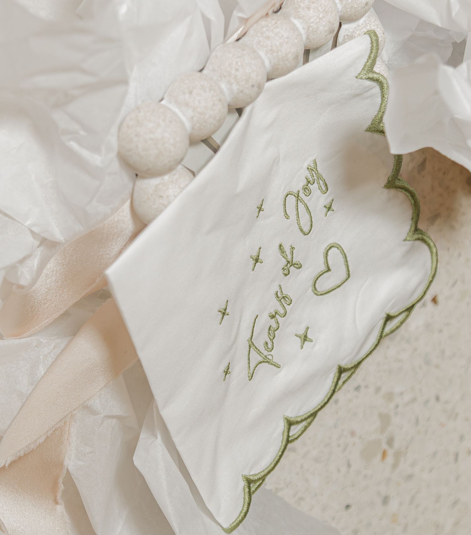 White embroidered napkin reading Tears of Joy with green scalloped edges, displayed among tissue paper and wooden beads.