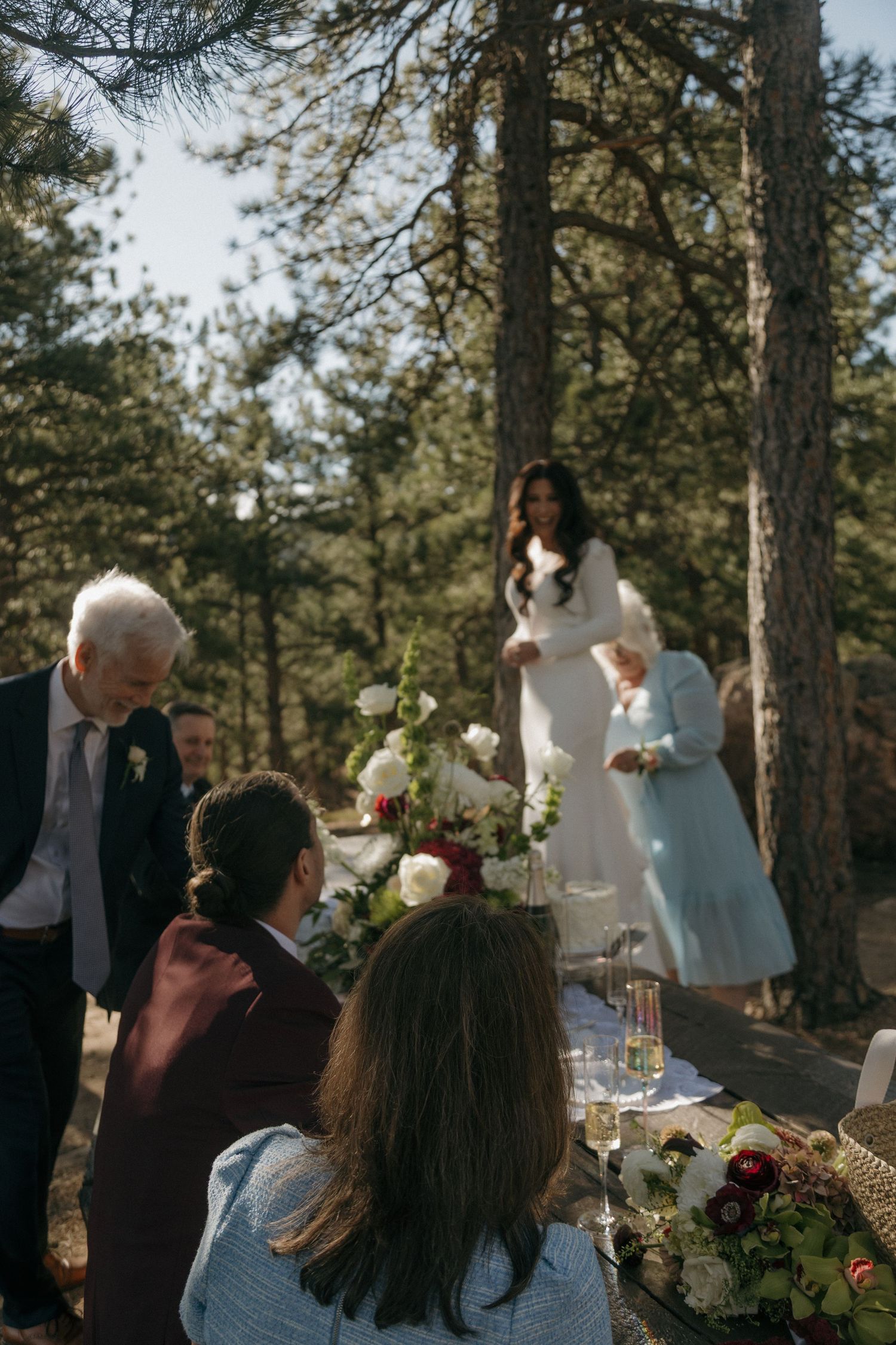 Outdoor woodland wedding reception with guests gathered around floral table as bride approaches in white gown.