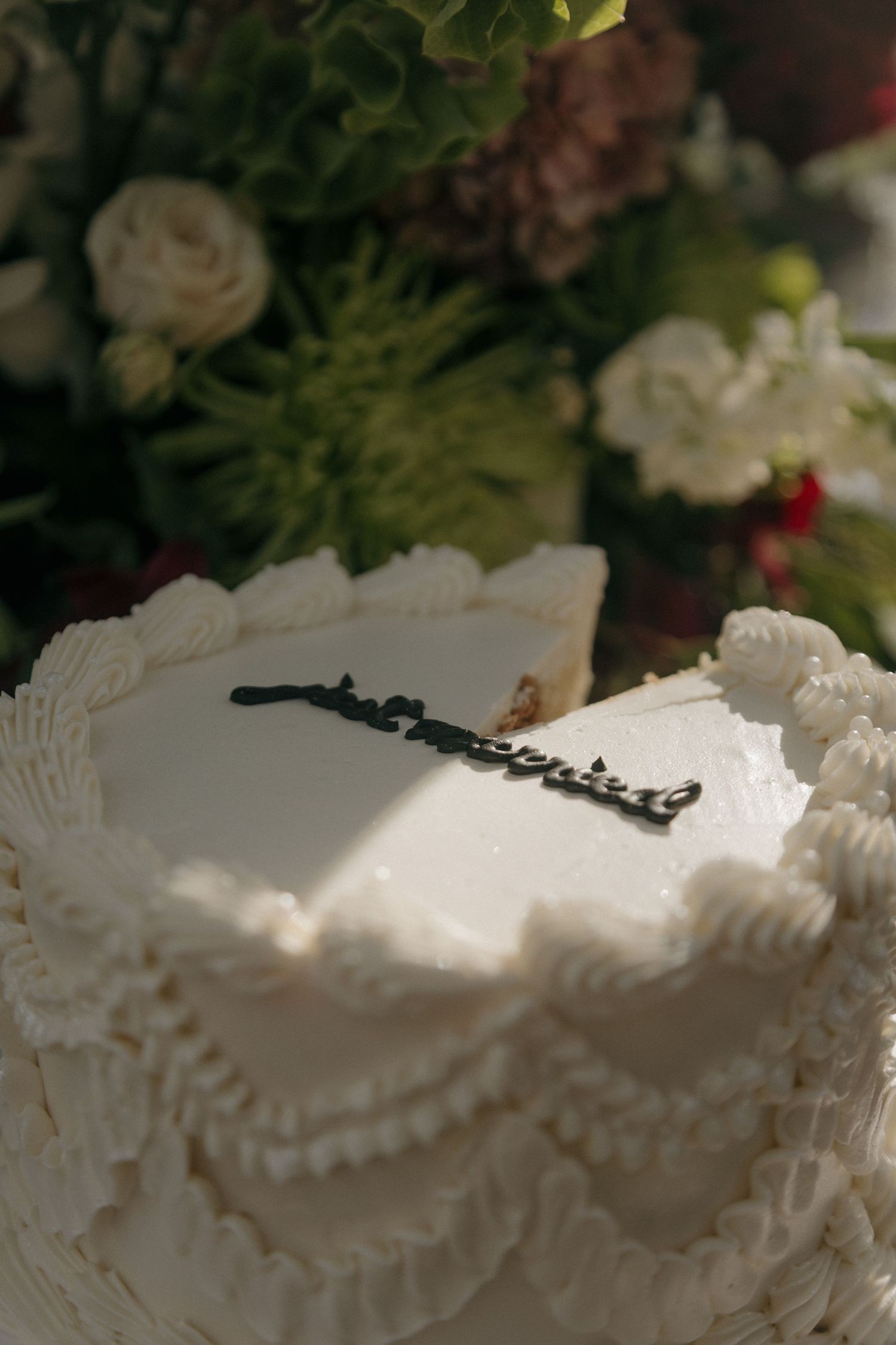 Elegant white wedding cake with ruffled frosting and decorative topper surrounded by lush floral arrangement.