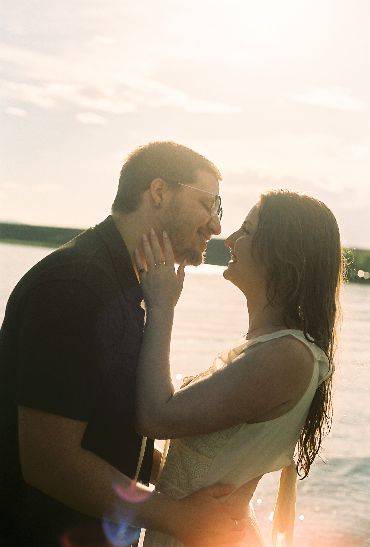 Couple about to kiss in bright sunlight by a lake, backlit by warm golden sun flare.
