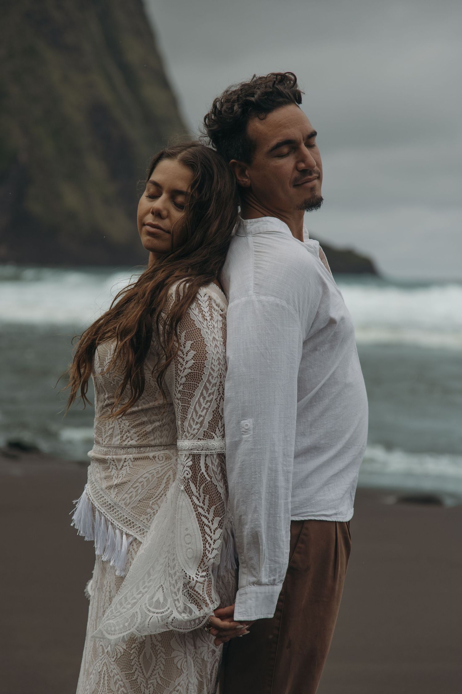 Wedding couple stands back-to-back on black sand beach with rocky sea cliffs in background.