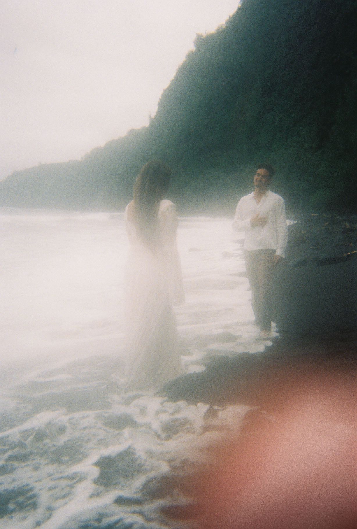 Dreamy film photo of bride in white gown and groom standing in ocean waves on a misty beach.