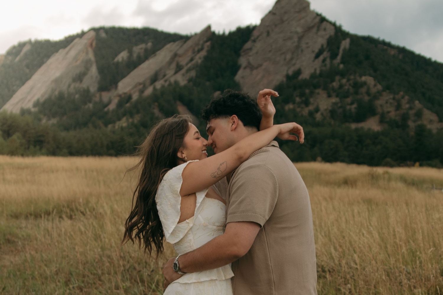 Romantic couple sharing a kiss in golden meadow with stunning rocky mountain flatirons behind them.