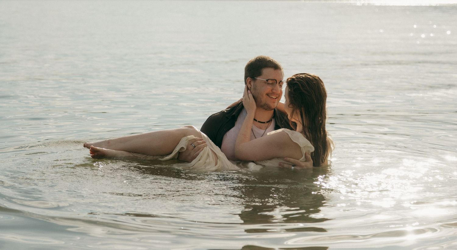 Couple embracing and laughing together while lying in shallow lake water during a playful engagement shoot.
