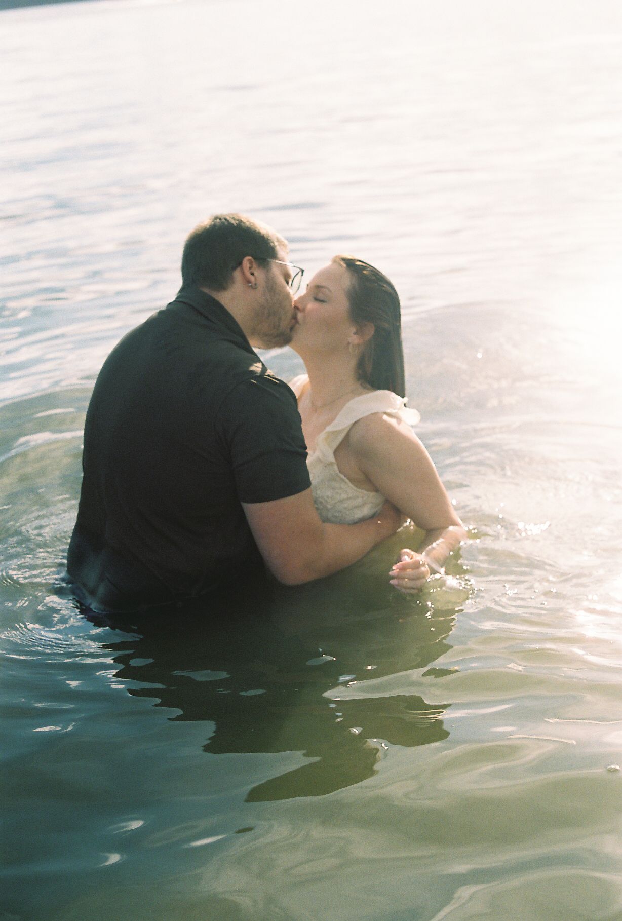 Couple sharing a passionate kiss while standing waist-deep in a glistening sunlit lake.