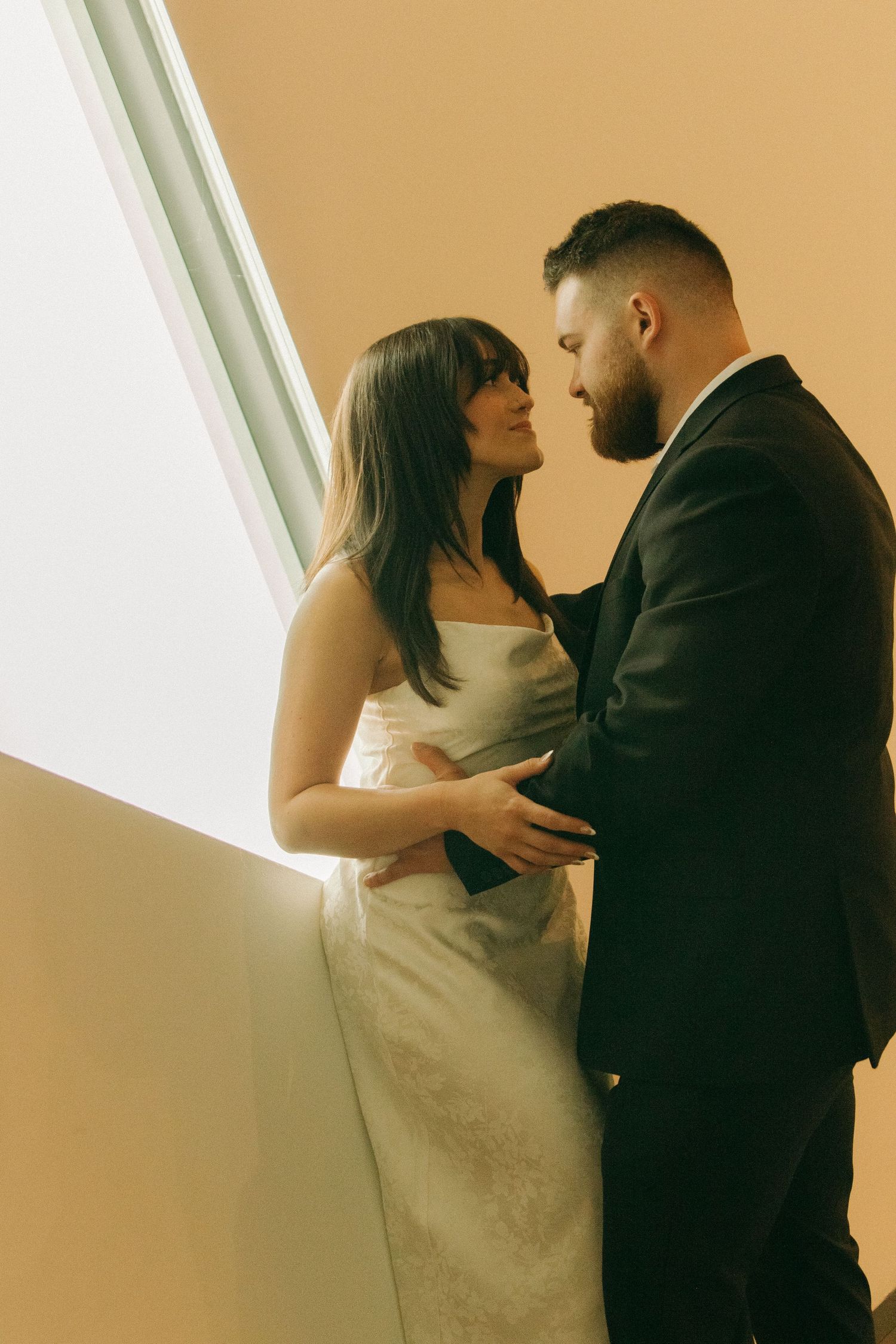 Couple embracing near bright window in formal attire.