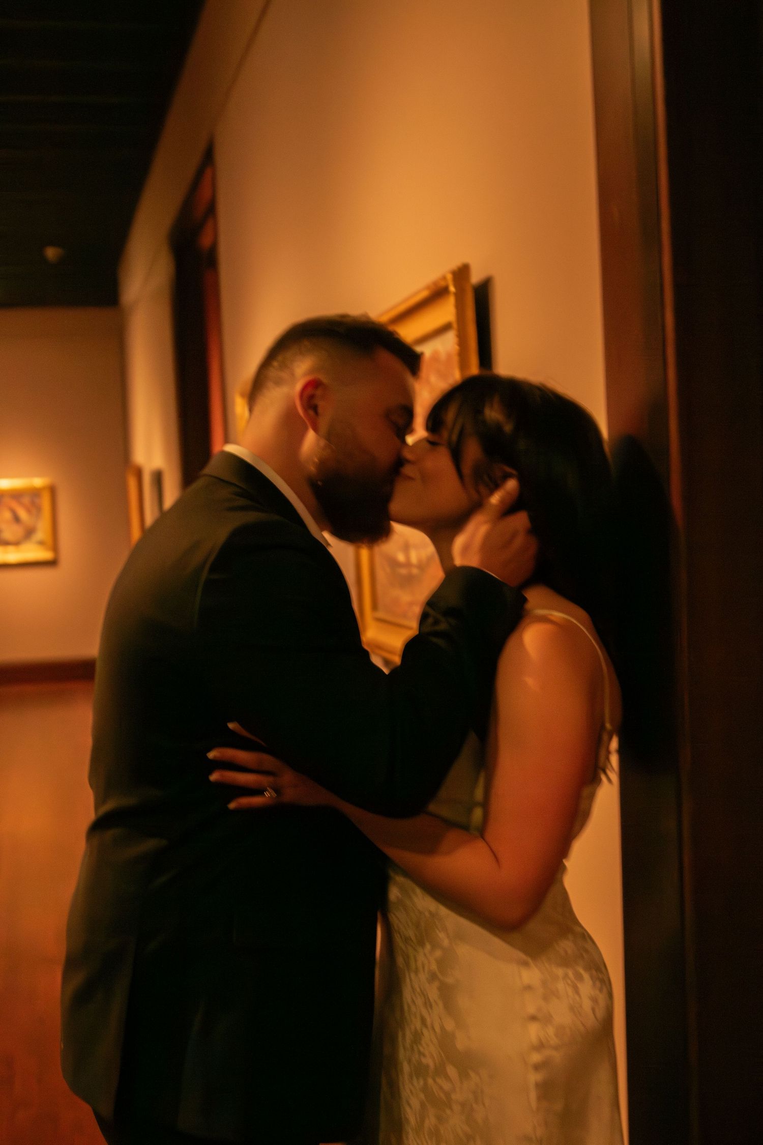 A couple shares tender moments in a warmly lit gallery hallway with artwork on display.