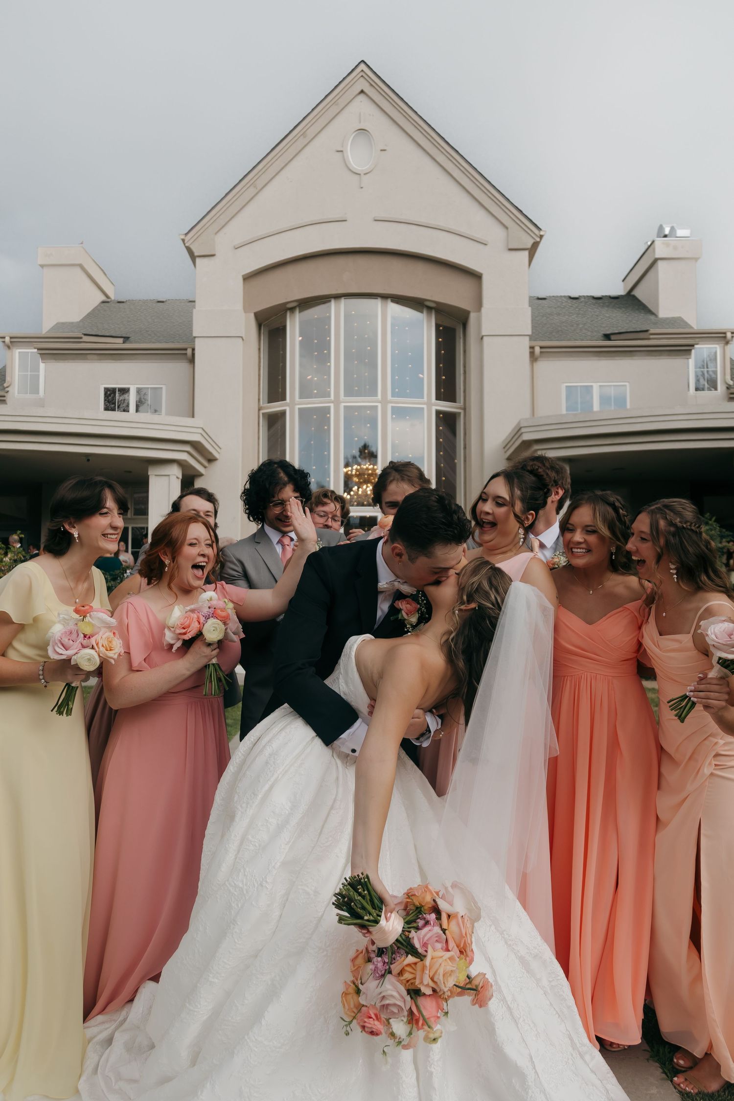 Bride and groom share a romantic dip kiss surrounded by cheering bridesmaids in colorful pastel gowns outside an elegant venue.