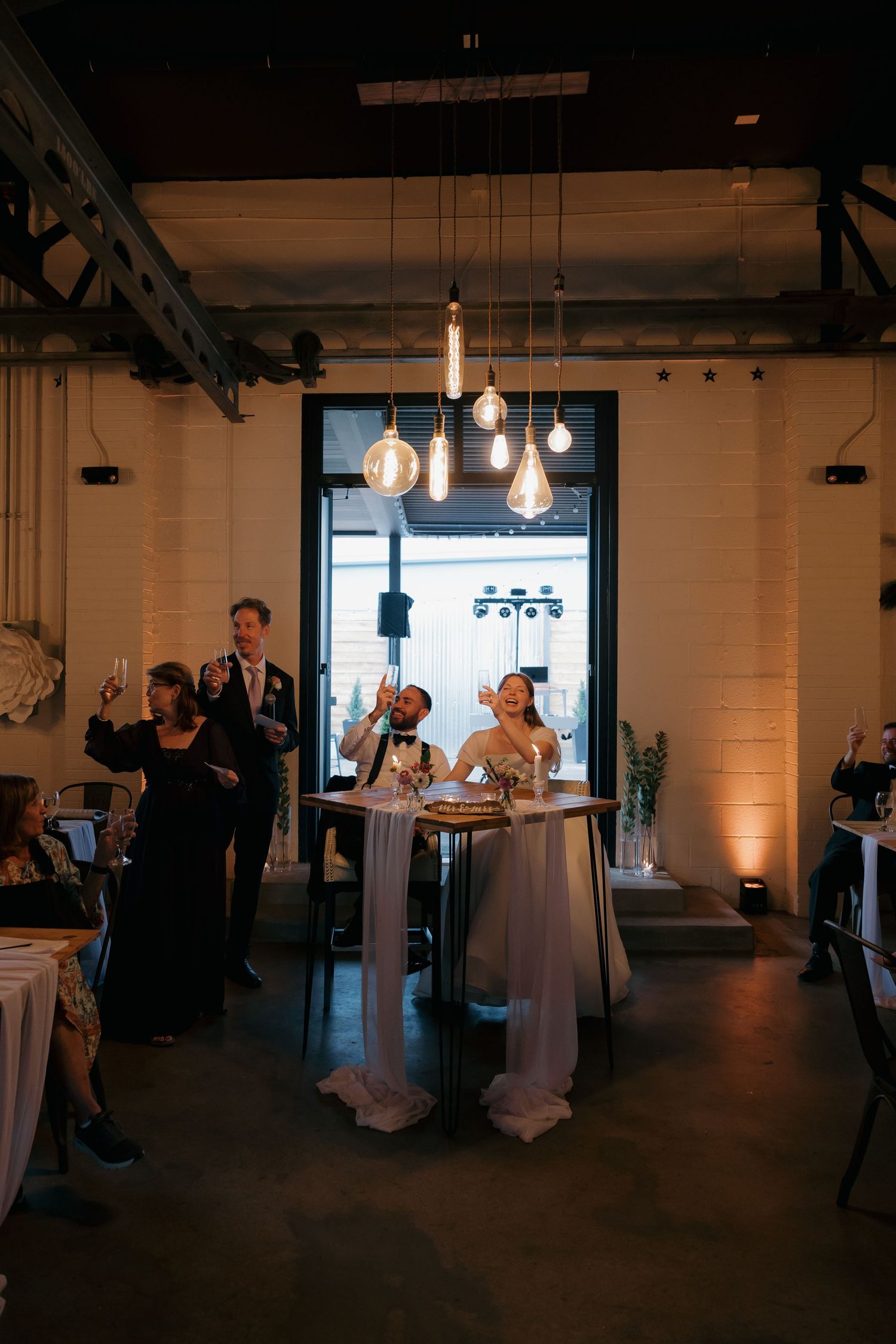 Bride and groom laugh at sweetheart table under hanging Edison bulb lights at industrial wedding venue.