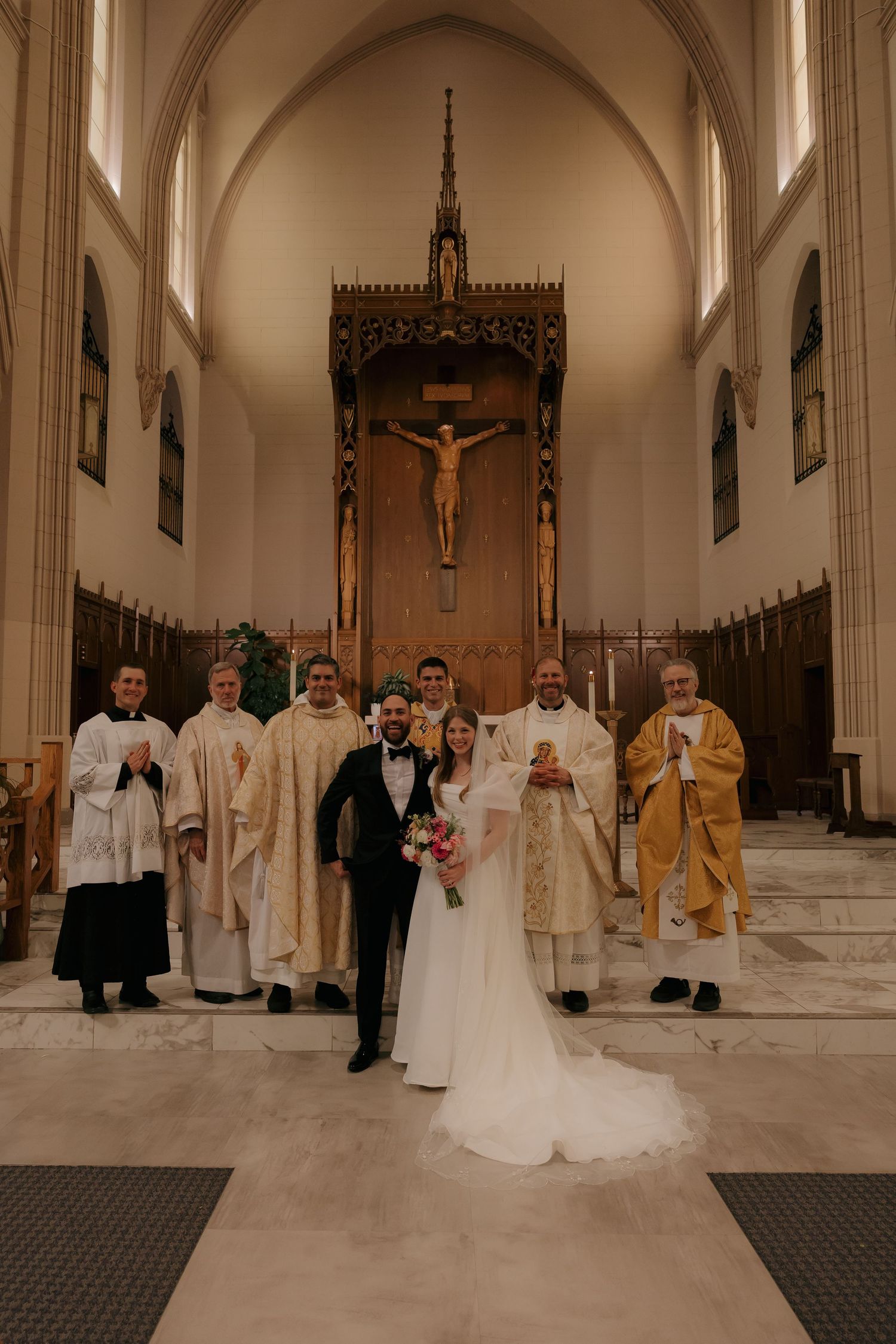 Newlyweds pose with priests at church altar beneath crucifix after wedding ceremony.
