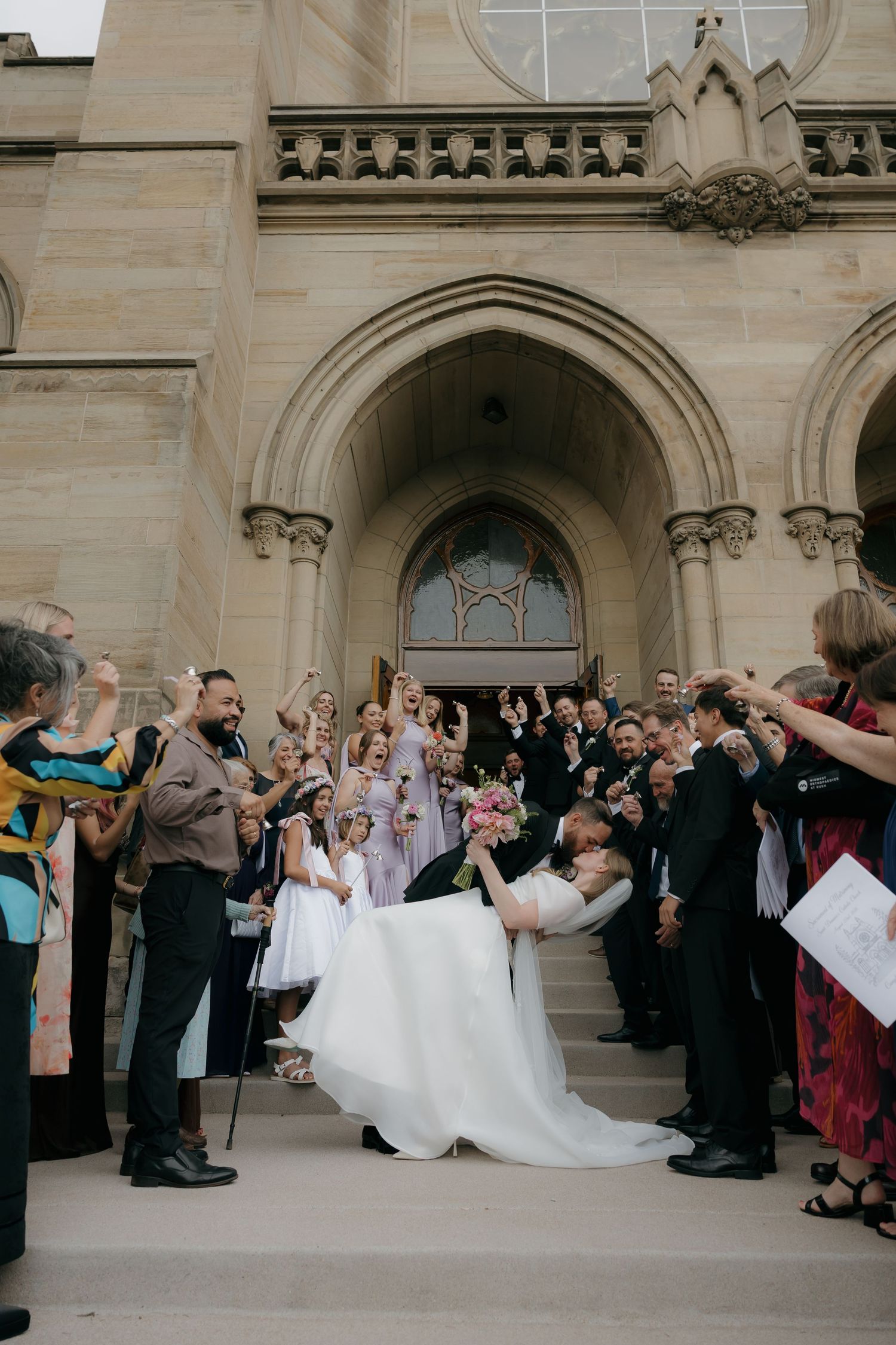 Groom dips bride outside a Gothic stone church as wedding guests cheer on the steps.