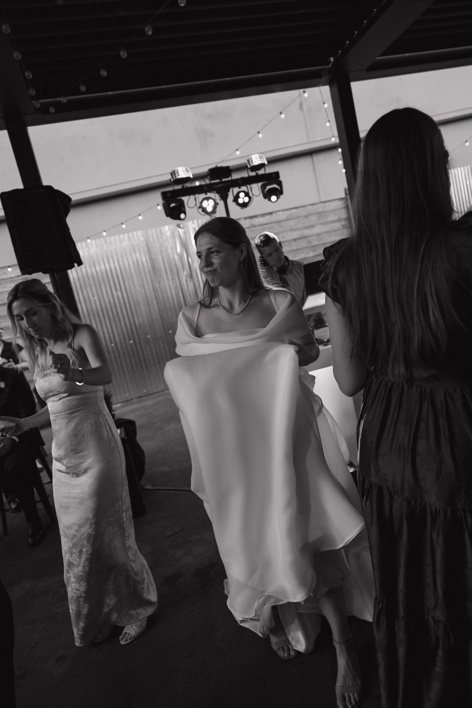 Black and white photo of barefoot bride dancing with guests at outdoor wedding reception under string lights.