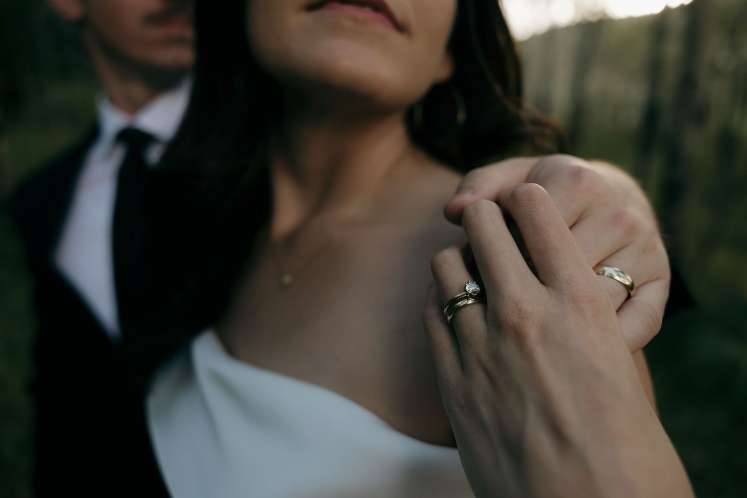 Close-up of bride's engagement ring and wedding band as groom's hand rests on her shoulder.