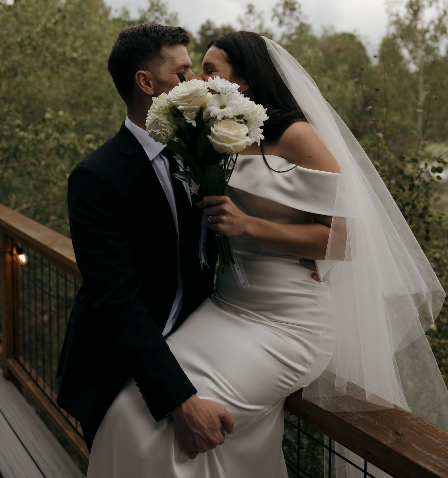 Bride and groom sharing romantic moment on wooden deck, bride holding white flower bouquet.
