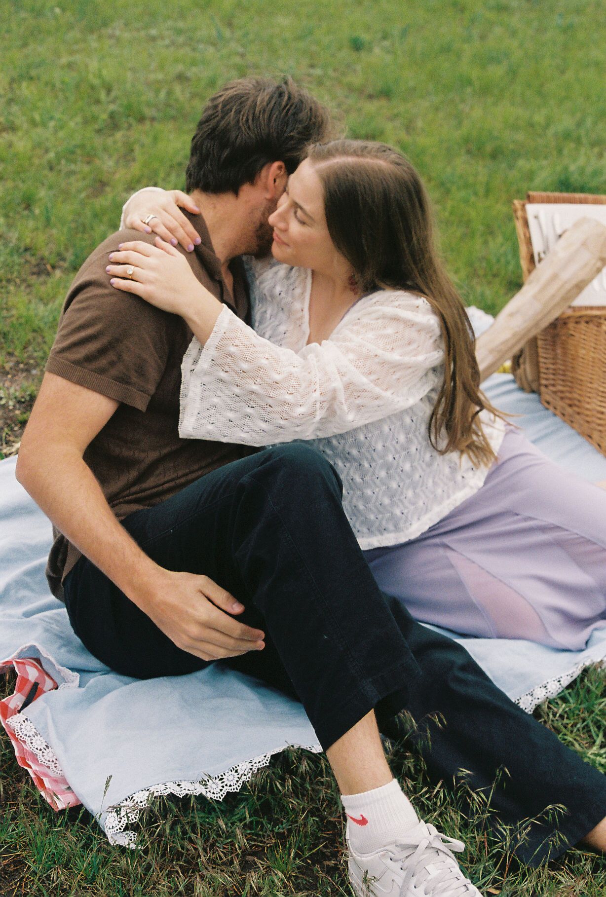Couple sharing a tender kiss while sitting on a picnic blanket outdoors with a wicker basket nearby.