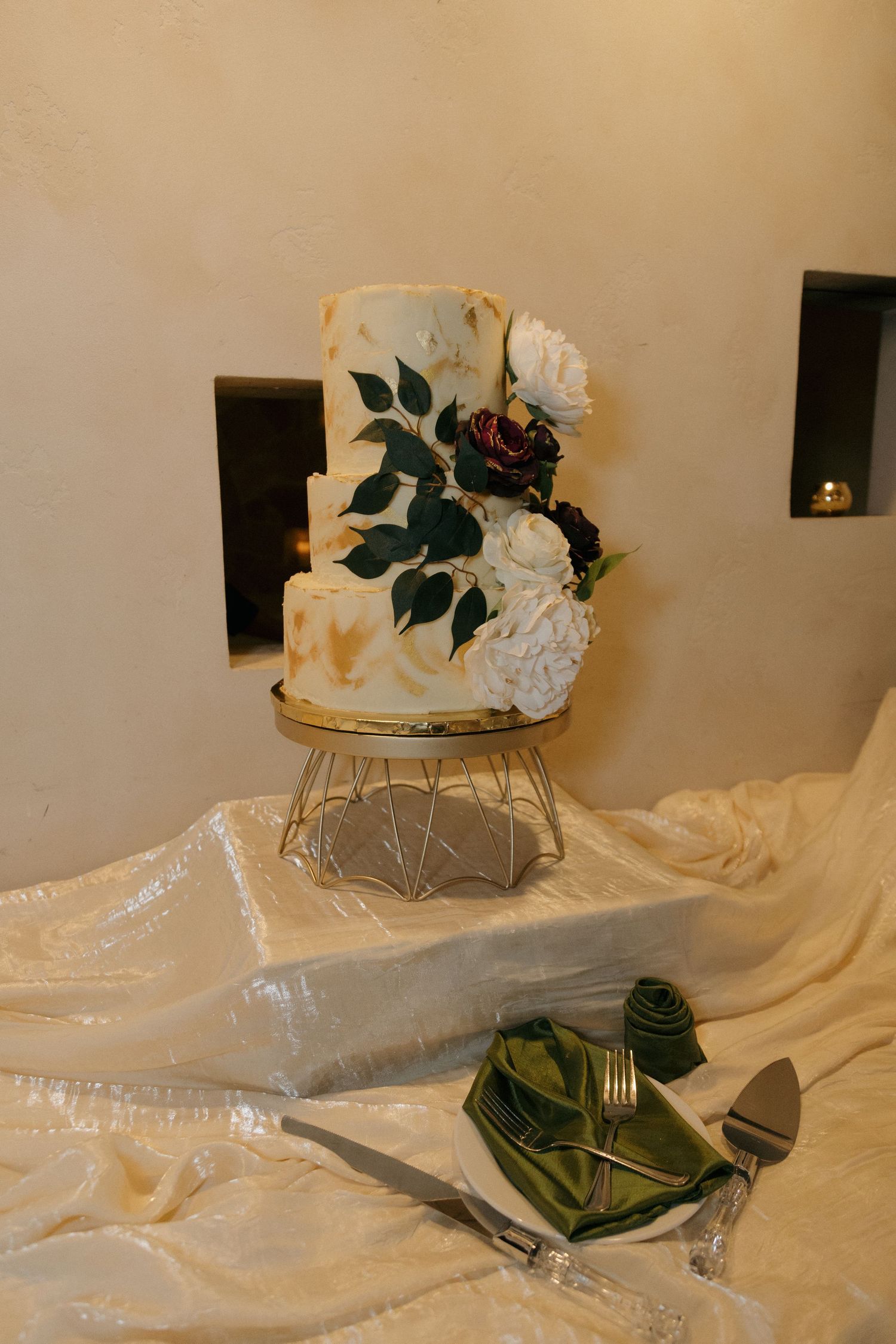 Three-tier white wedding cake with gold accents and floral decorations on gold wire cake stand.